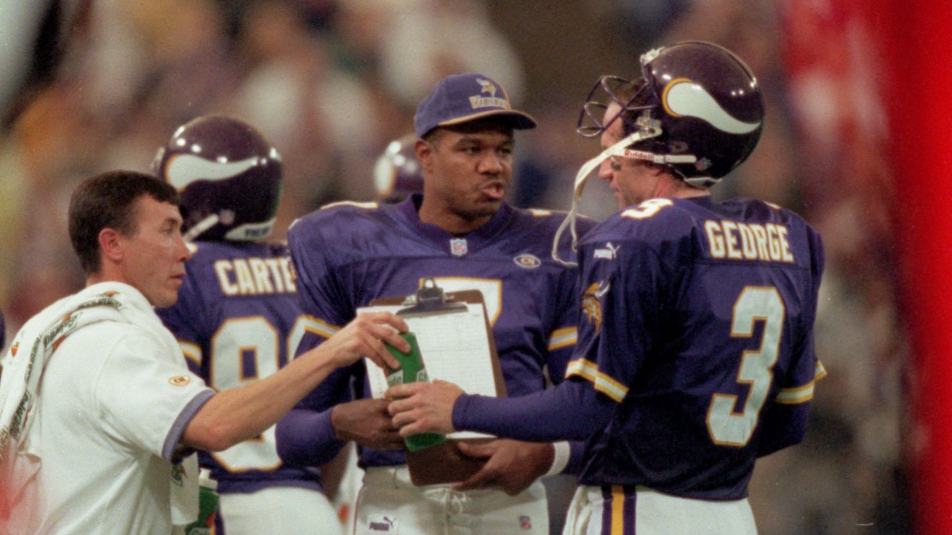 Randall Cunningham, wearing a purple Vikings hat and jersey, talks to Jeff George, also wearing a purple jersey. A coach wearing a white polo shirt is talking to them. 