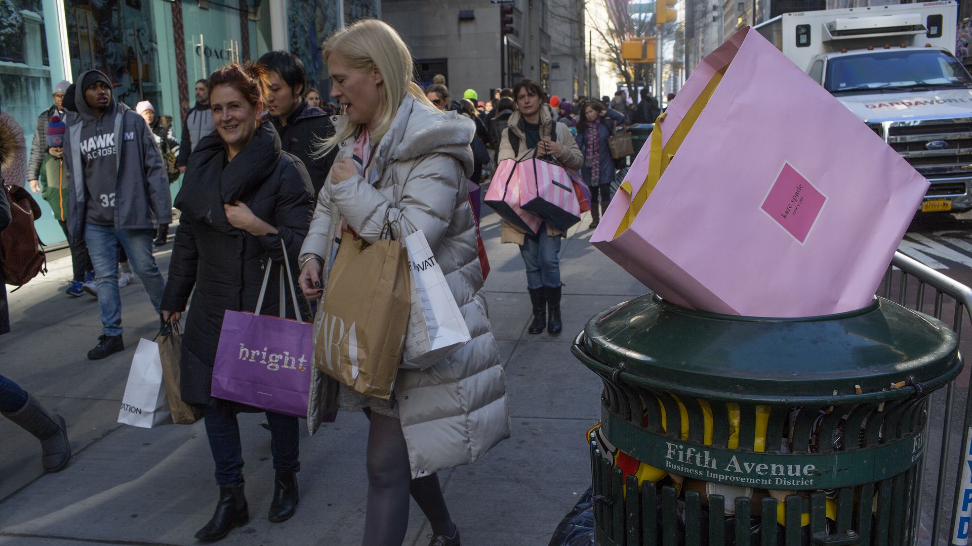 In this image, a pink shopping bag sits on top of a trash can while holiday shoppers walk down the street
