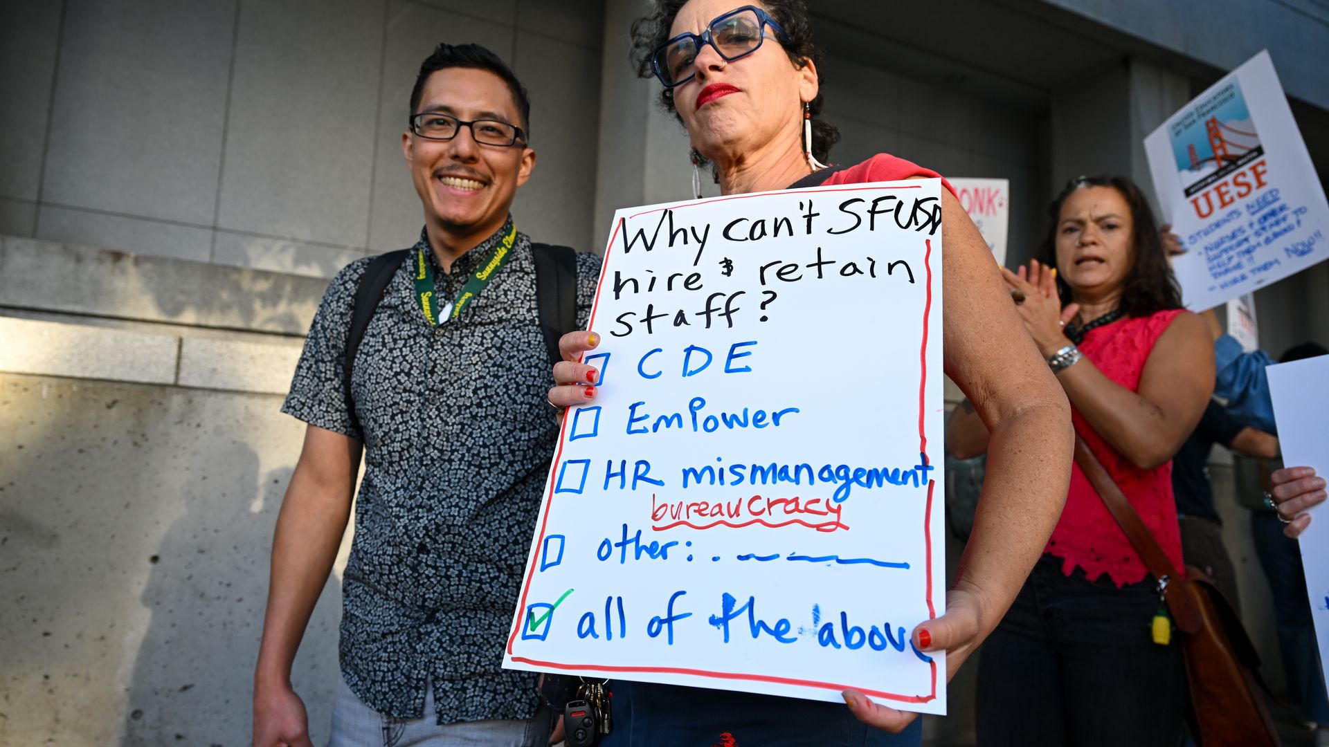 Woman with curly hair and blue glasses holds a protest sign asking why SFUSD can't hire and retain staff, listing reasons including HR mismanagement and bureaucracy, with a man smiling beside her.