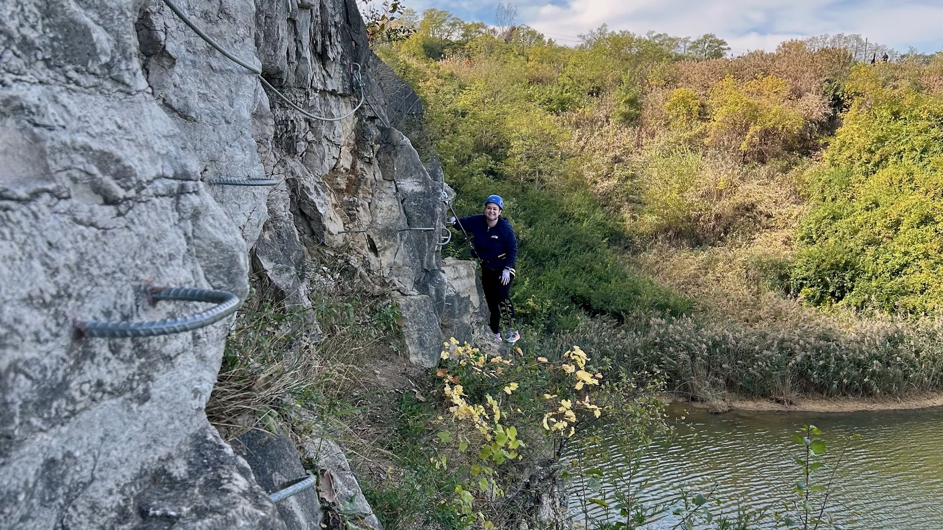 Alissa, in a blue helmet and dark clothes, climbing a rocky cliff with metal steps, near a body of water surrounded by green and brown foliage under a partly cloudy sky.