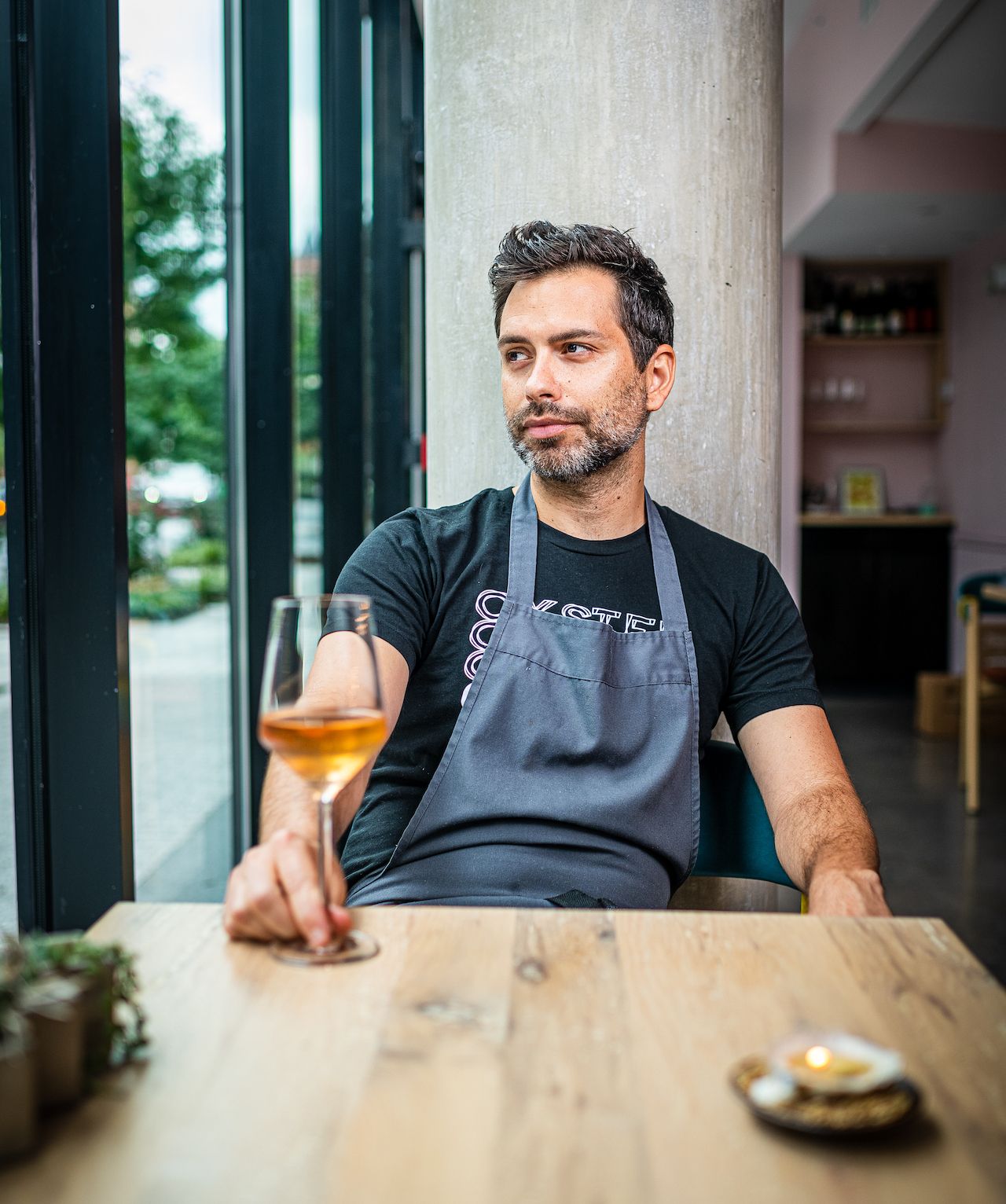D.C. chef Rob Rubba in a black t-shirt and blue apron sitting at a table with a glass . 