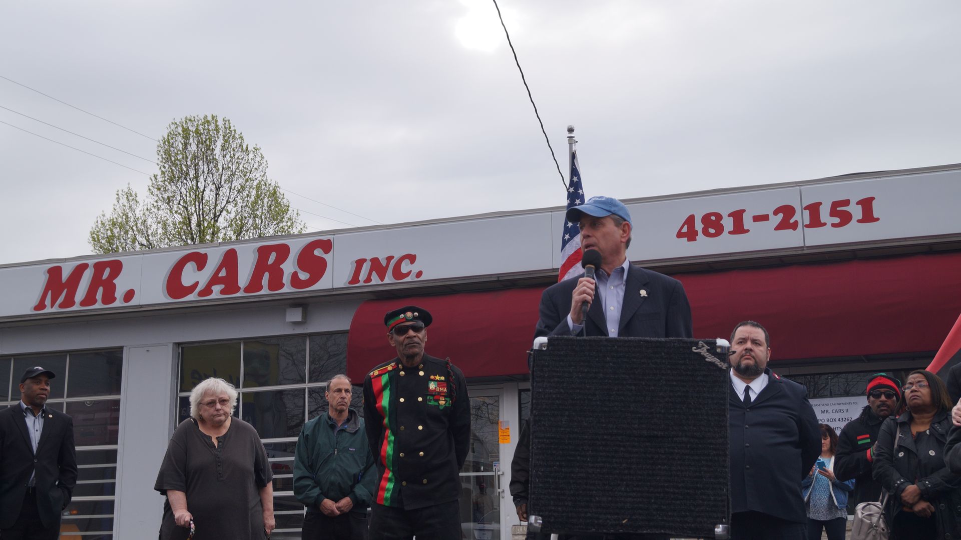 A man in a blue cap speaks into a microphone at a podium outside Mr. Cars Inc., with several people standing behind him, including a man in a black, red, and green uniform and an American flag.