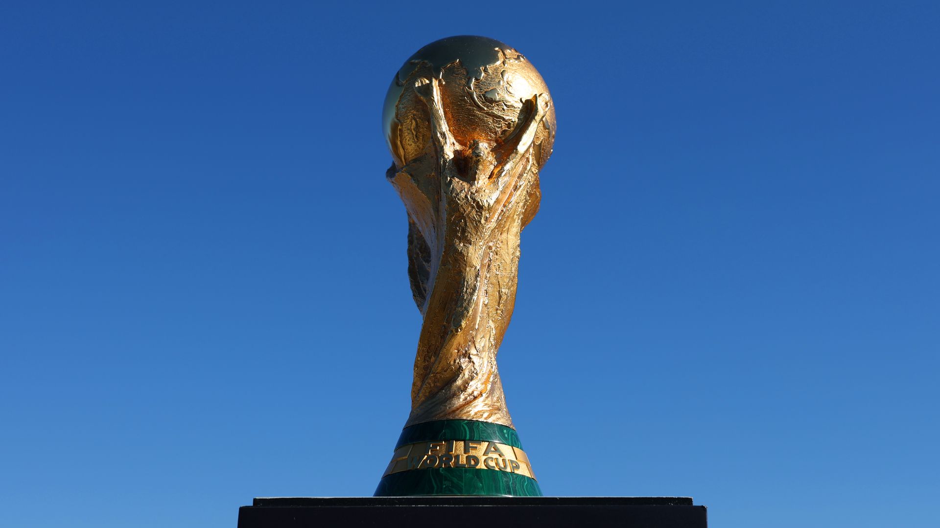 Golden FIFA World Cup trophy with a globe on top, featuring a figure holding it up against a clear blue sky.