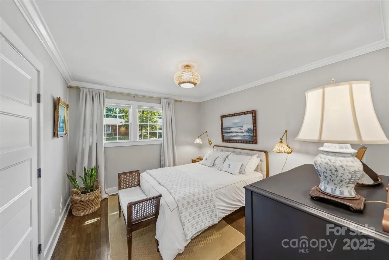 Bright bedroom with white walls, bed with white linens, wooden headboard and bench, large window with white curtains, wall lamps, framed ocean painting, wooden floor, and a lamp on dark dresser.
