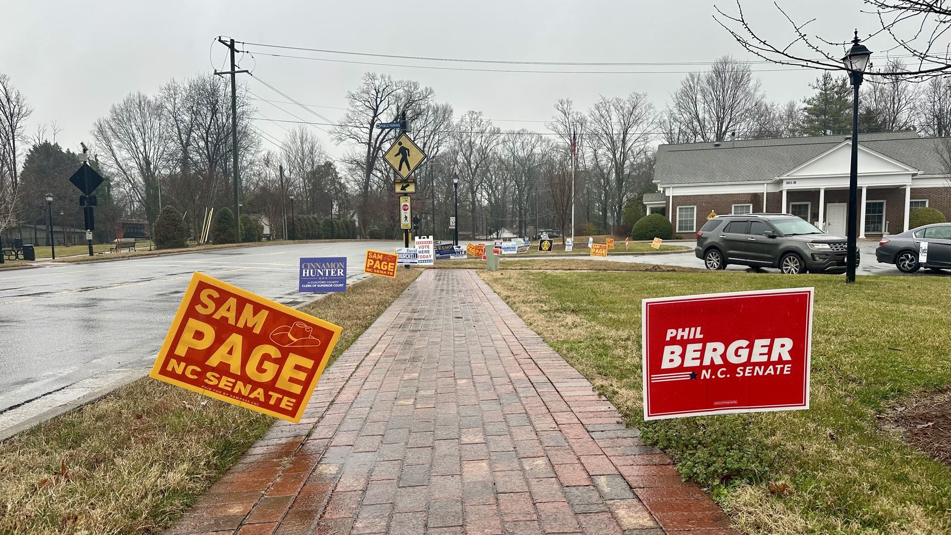 Brick sidewalk on a rainy day with numerous political campaign signs on grass, including red and yellow "Sam Page NC Senate" and red "Phil Berger N.C. Senate" signs, in a suburban area.