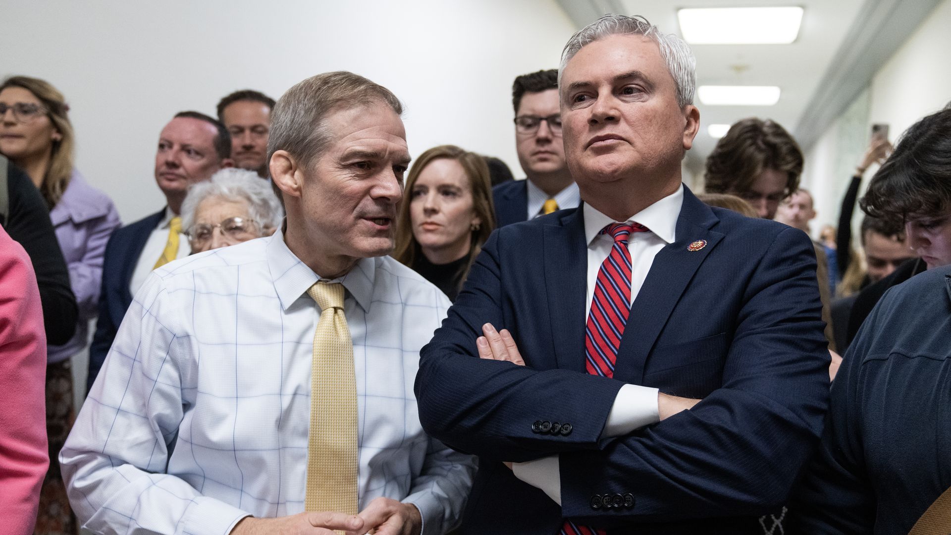 House Judiciary Committee Chair Jim Jordan wearing a white shirt and gold tie and House Oversight Committee Chair James Comer wearing a blue jacket, white shirt and red and blue striped tie, surrounded by reporters and colleagues in a House office building hallway.