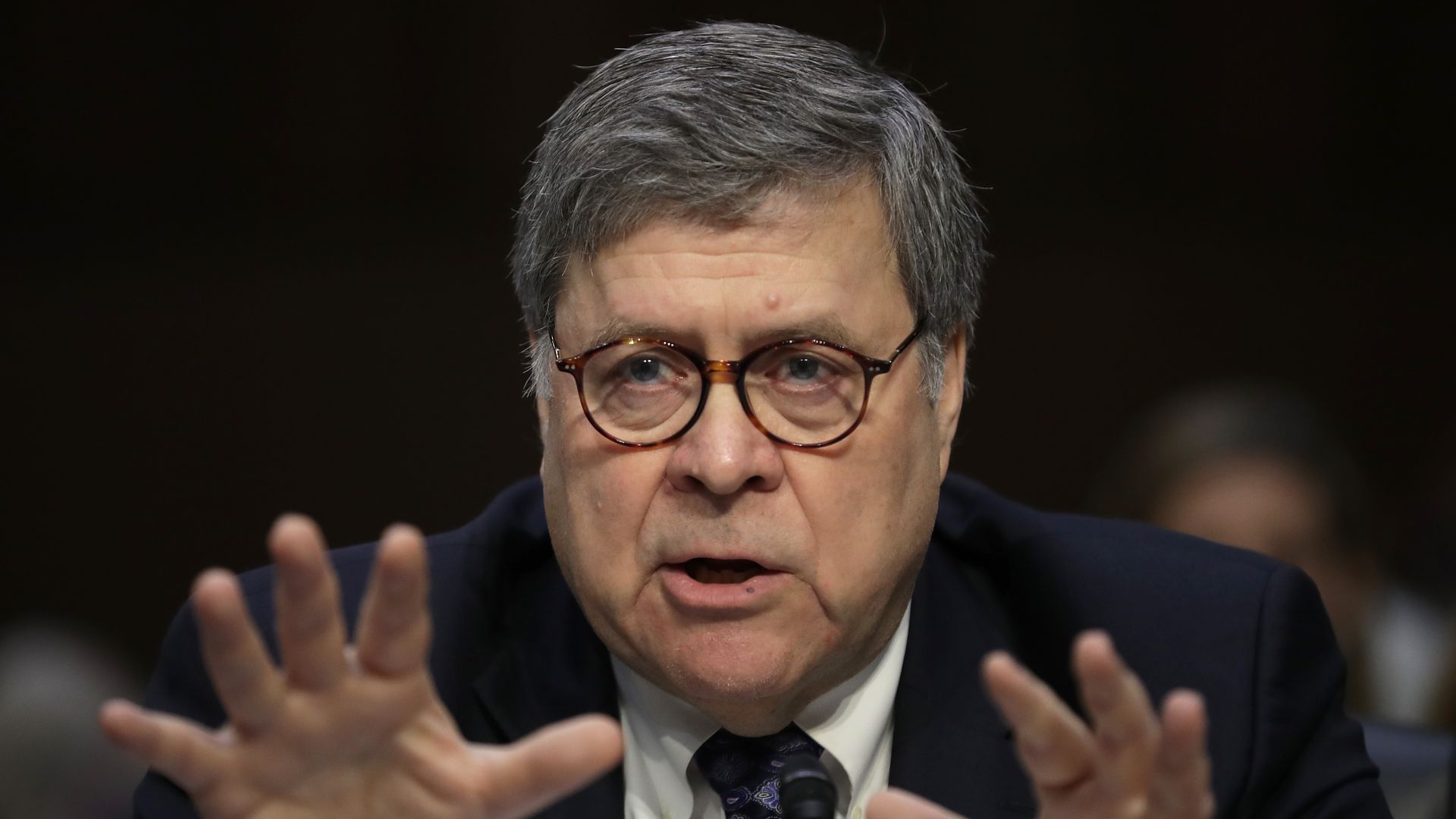 In this image, Barr speaks with both hands outstretched towards the camera. 