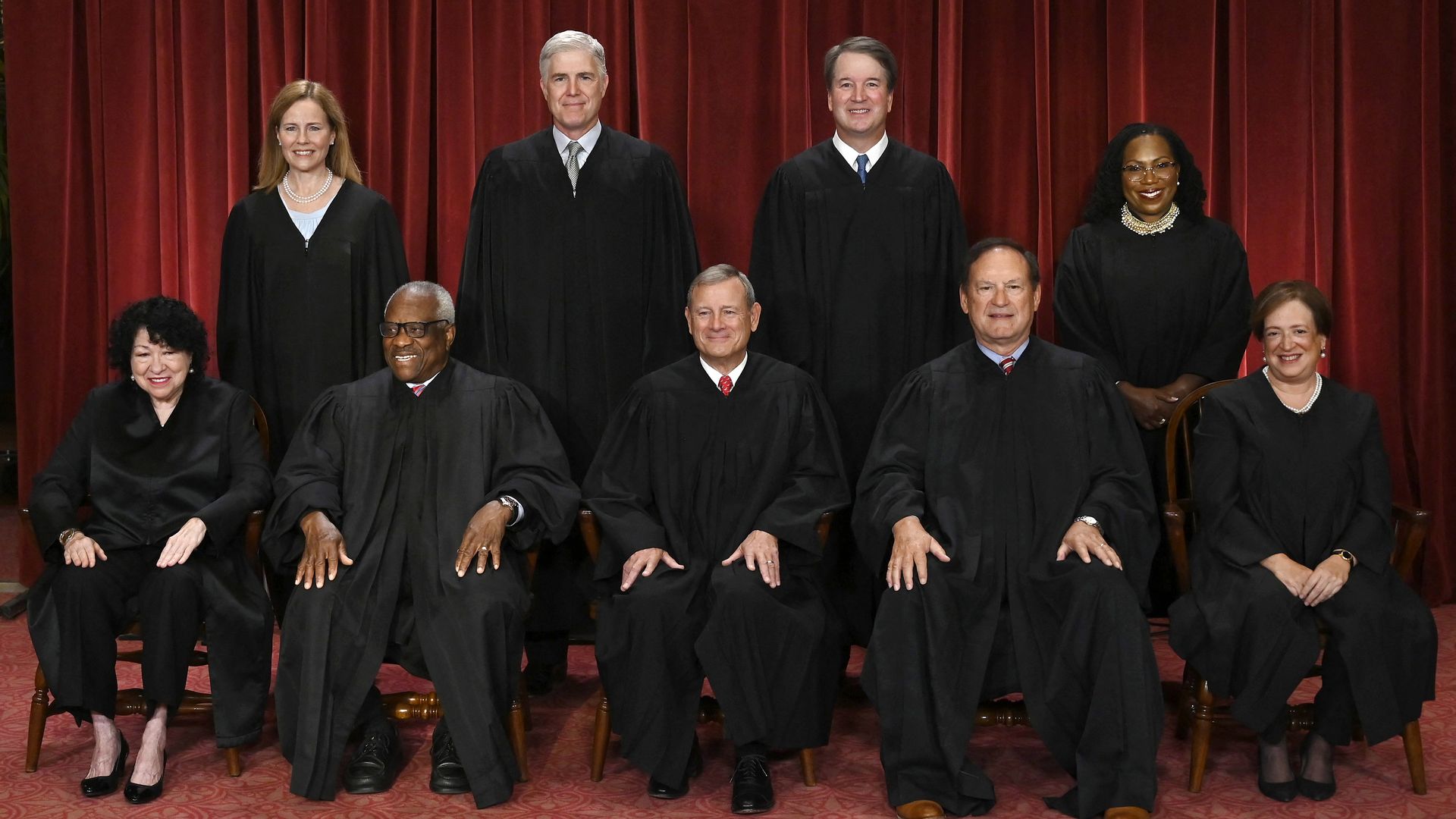 The Supreme Court justices in black robes posing on a stage with a deep red curtain, some standing and others seated, smiling for the photo.