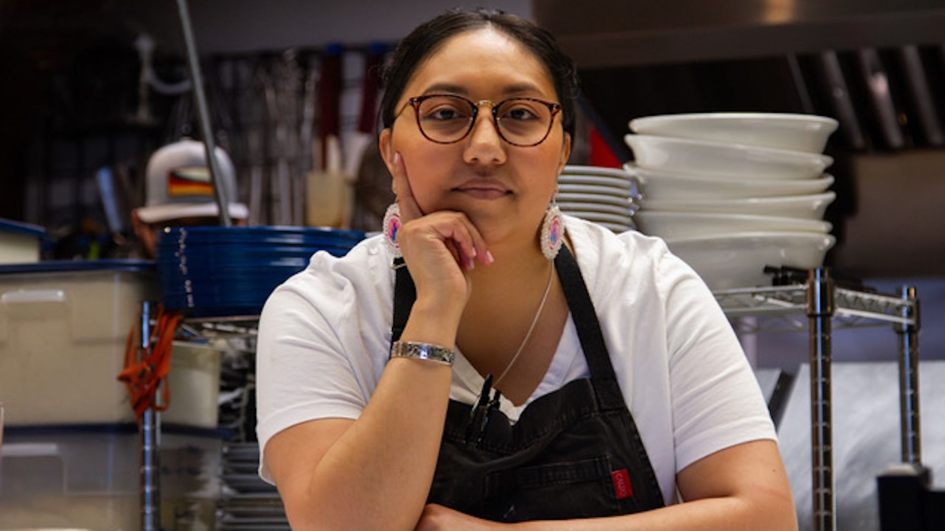 Woman wearing glasses, white shirt, and black apron with colorful earrings, sitting with hand on chin in a kitchen with stacked white plates and kitchen equipment.