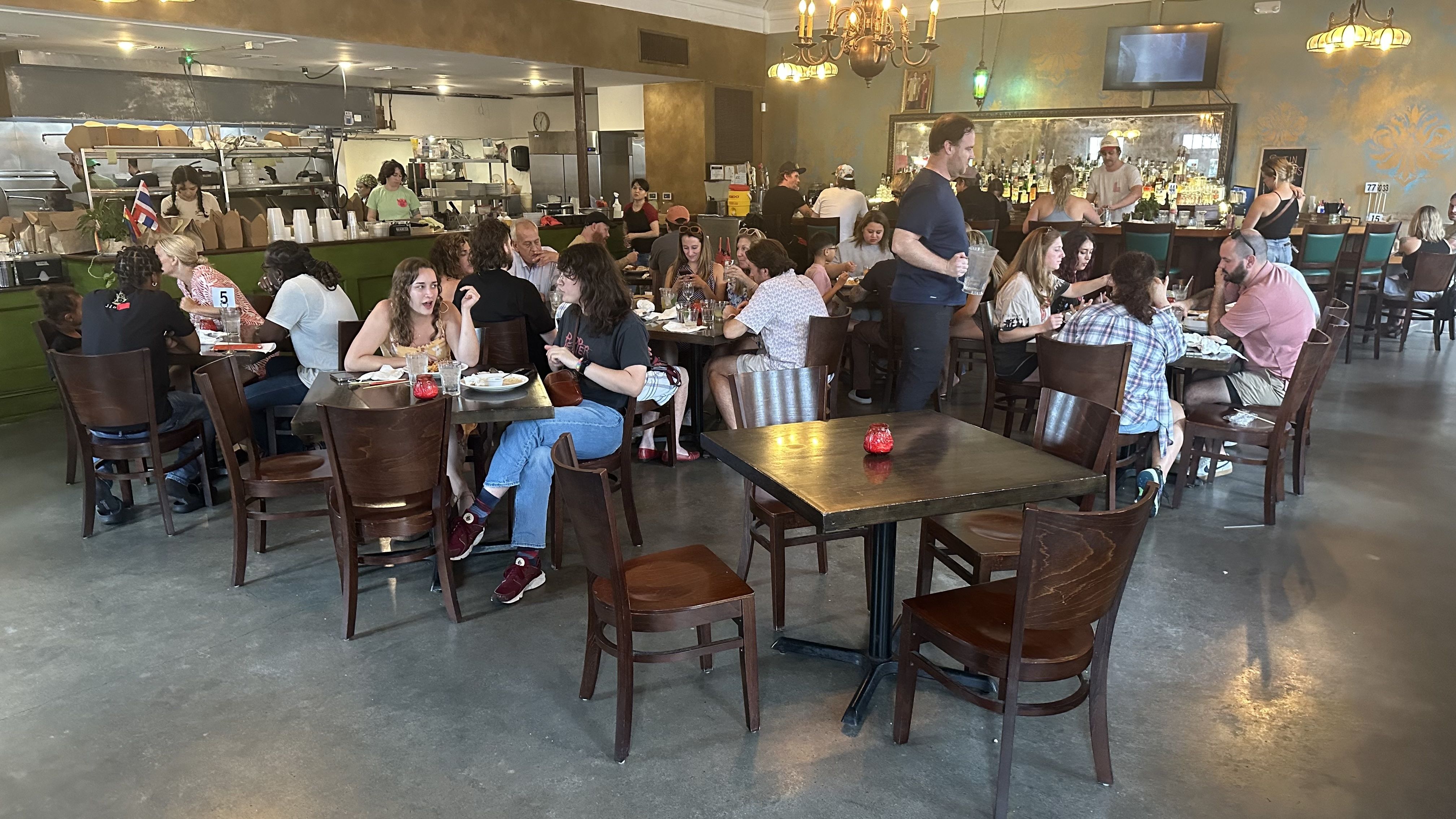 Busy restaurant interior with patrons seated at wooden tables, a green counter staffed by servers, chandeliers overhead, and a bartender behind the bar filled with bottles.