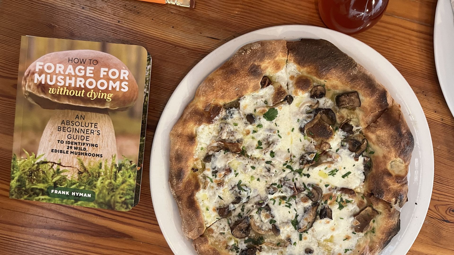 Top-down view of a wooden table with a mushroom pizza on a plate, a book titled "How to Forage for Mushrooms Without Dying", a pencil and a red spice oil carafe.