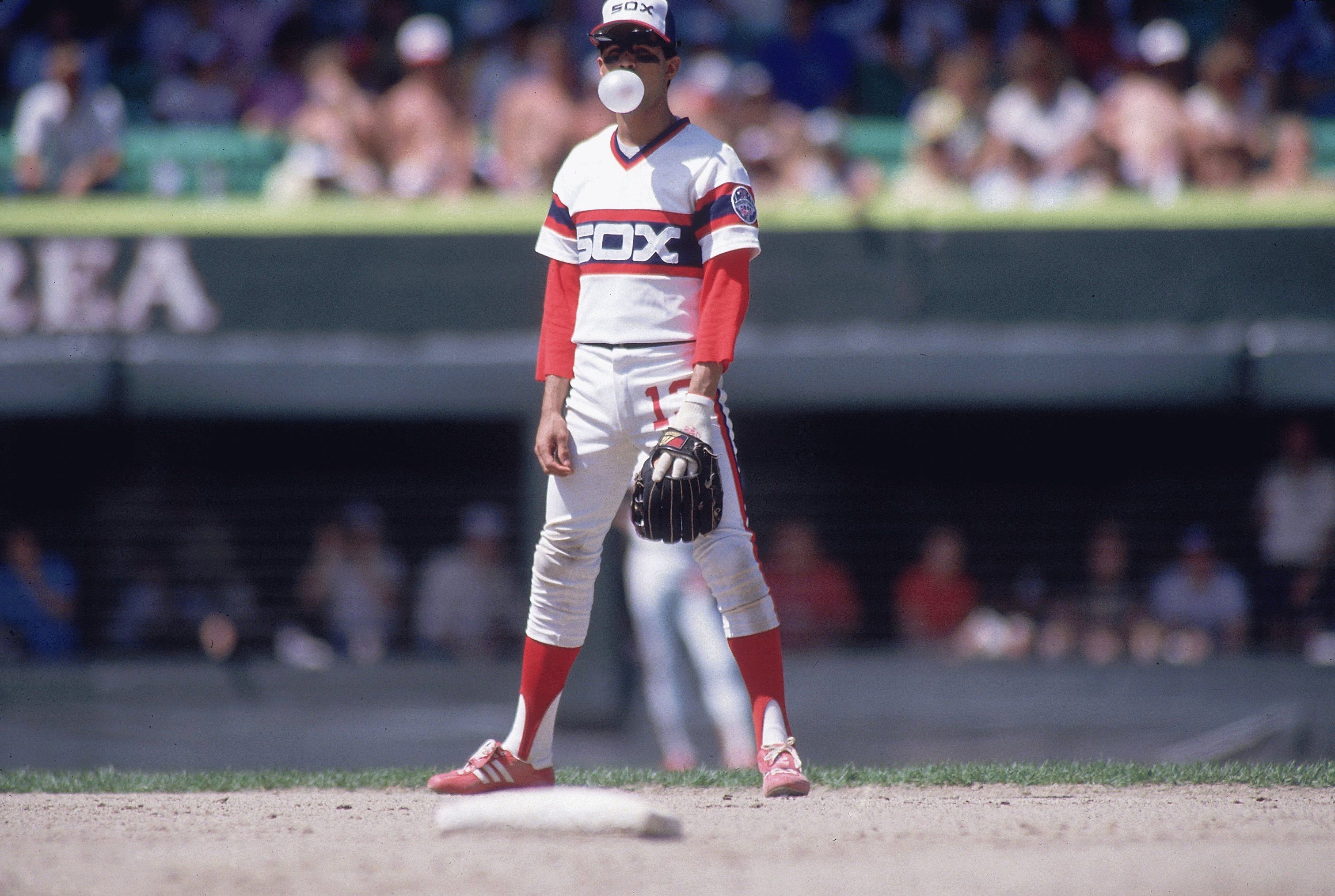 Baseball player in a White Sox uniform stands on the infield, blowing a bubble with gum. Glove in hand, red socks, cap on head; blurred crowd and a white base visible behind.