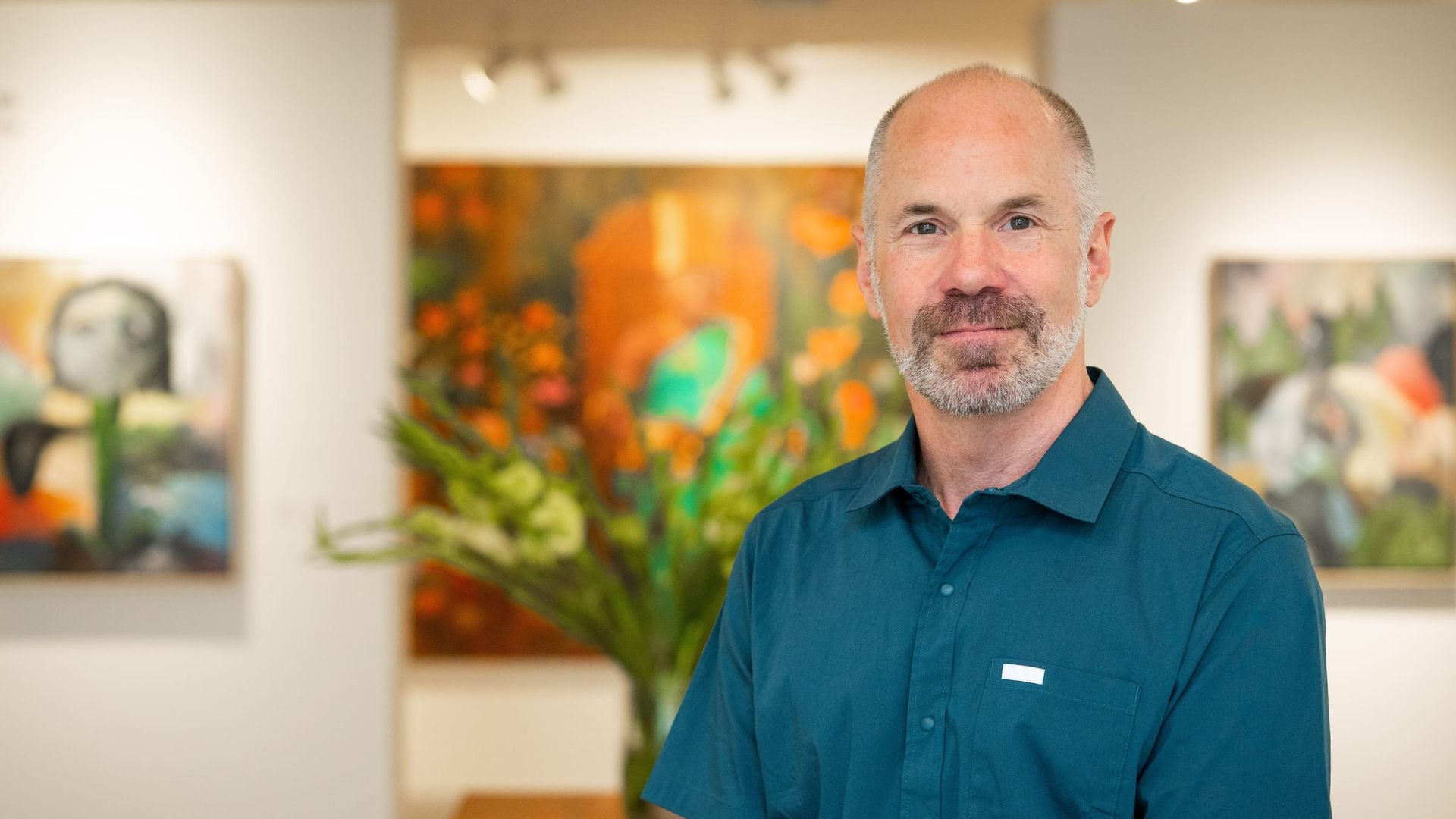A middle aged white man with a grey beard  in an art gallery with paintings and flowers in the background