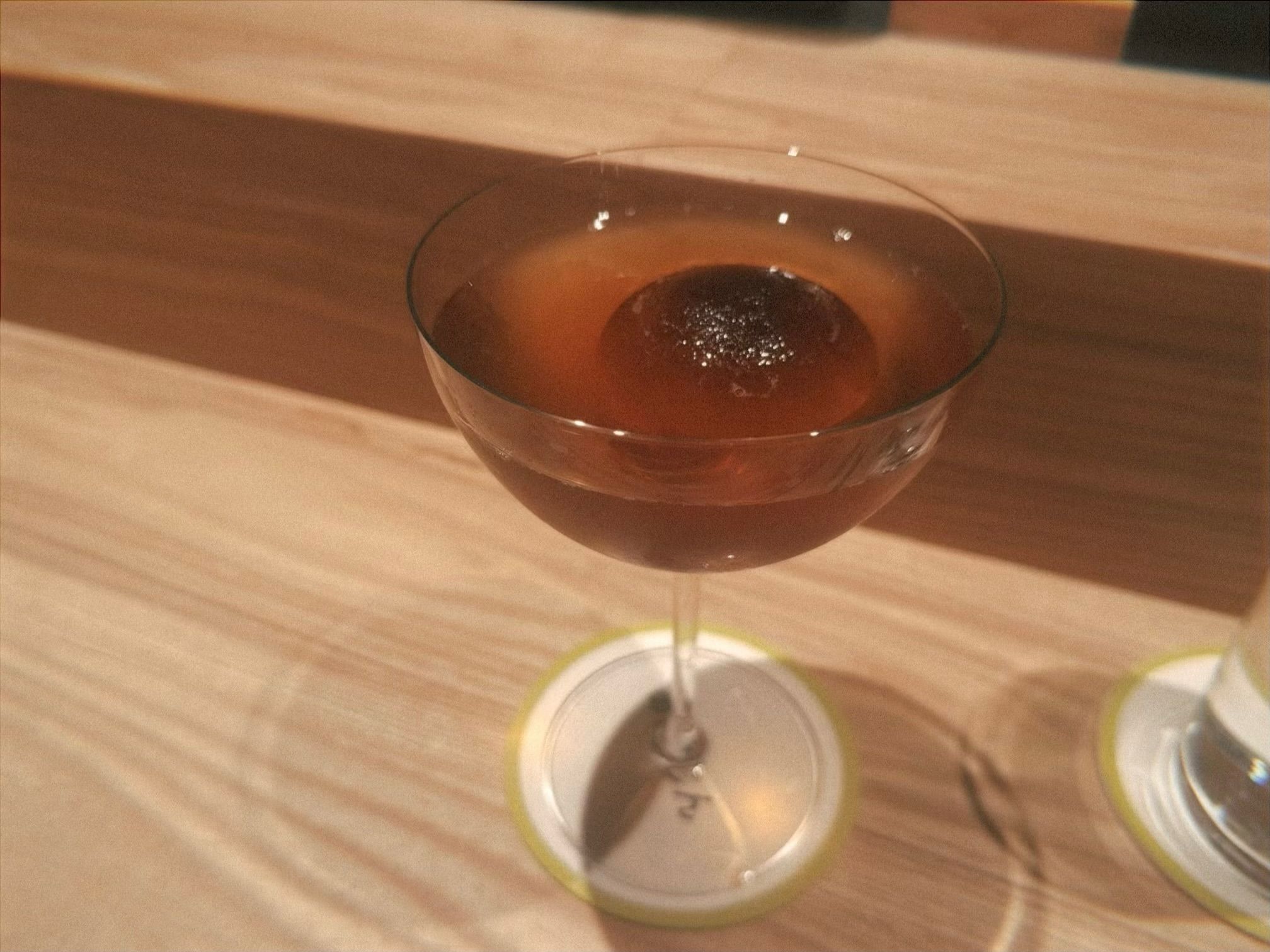Amber-colored cocktail in a coupe glass with a large round ice cube, placed on a white round coaster with a green edge on a wooden surface.
