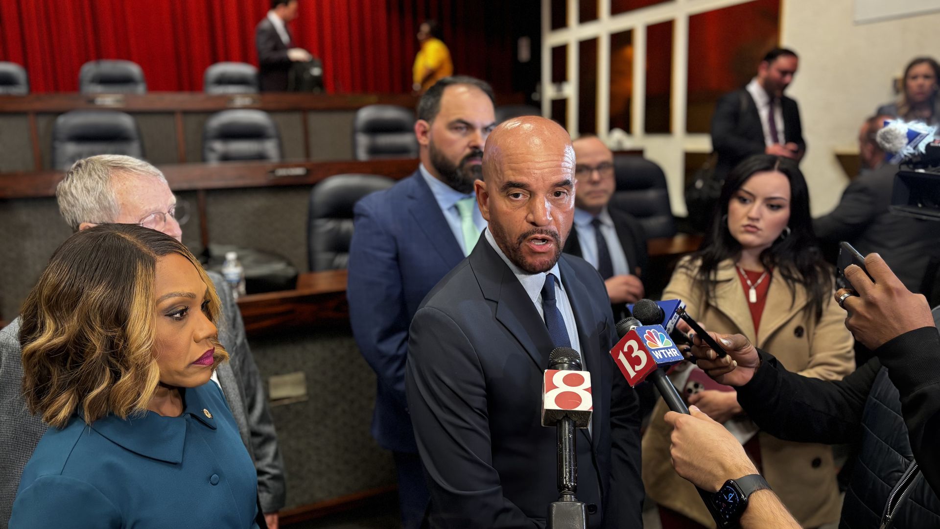 Bald man in a dark suit speaks to reporters at a press conference; microphones show "8" and "WTHR". A woman in teal stands left as other officials observe.