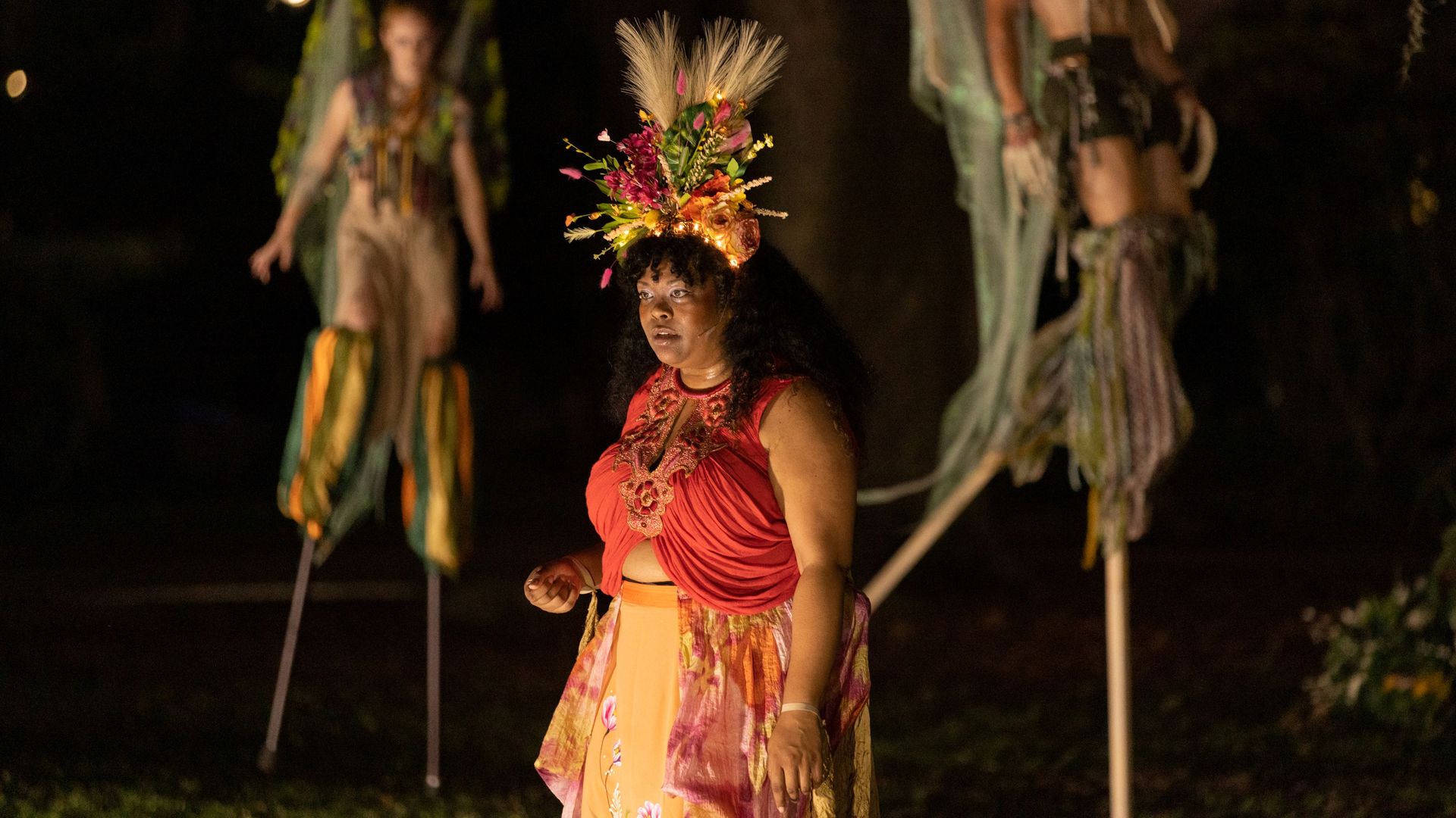 A trio of actors, each dressed whimsically, perform. One actor stands at center, staring toward an unseen audience, and two others approach her, dressed as fairies, on stilts.