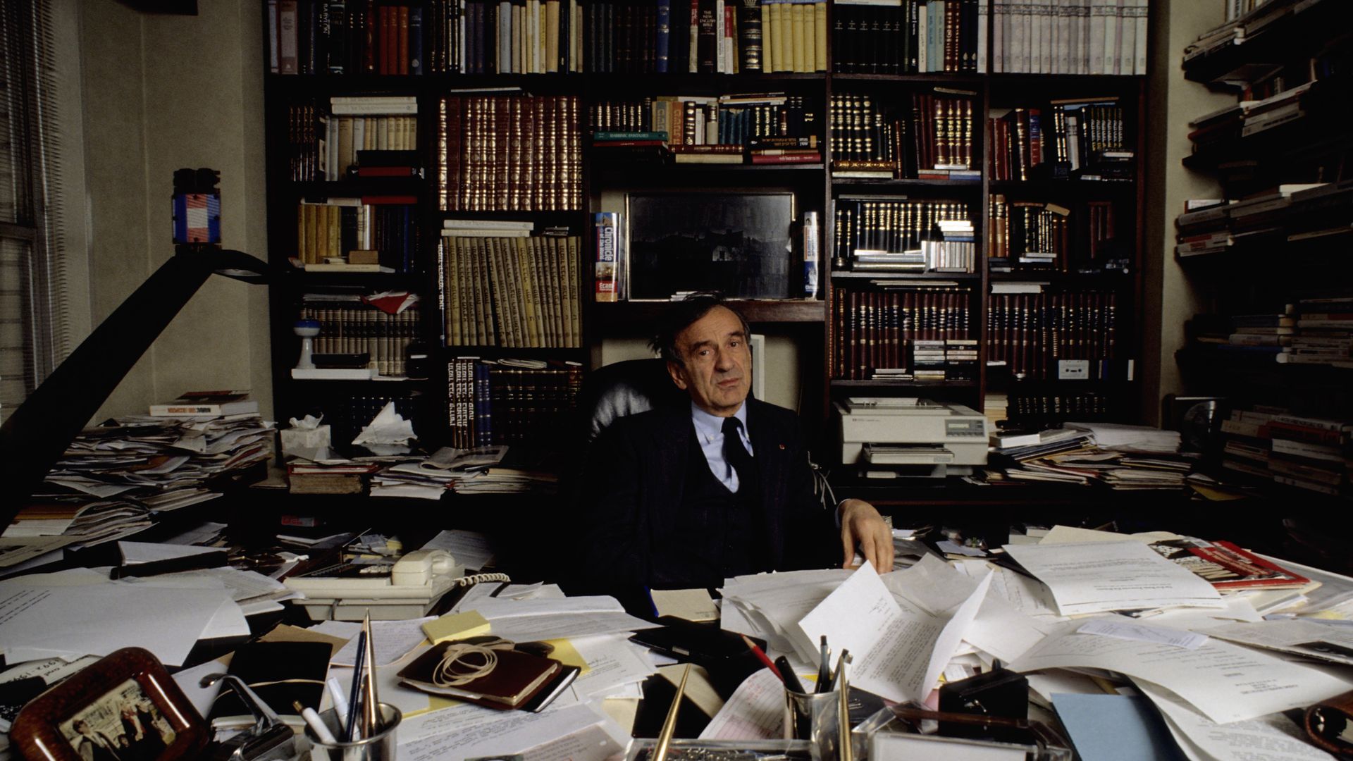 Elie Wiesel sits at a paper-strewn desk with bookshelves in the background 
