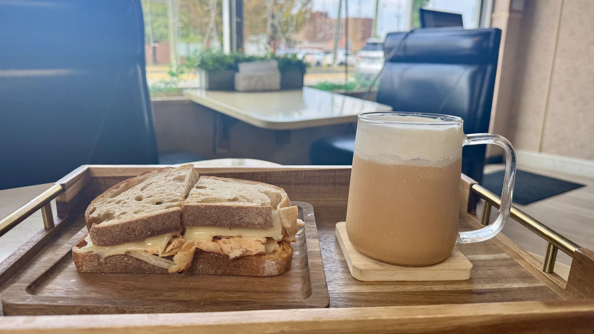 A wooden tray holds a buffalo chicken and cheese sandwich on rustic bread beside a frosted glass mug of iced tea with cold foam; in a cafe with black leather booth chairs and a table visible in the background.
