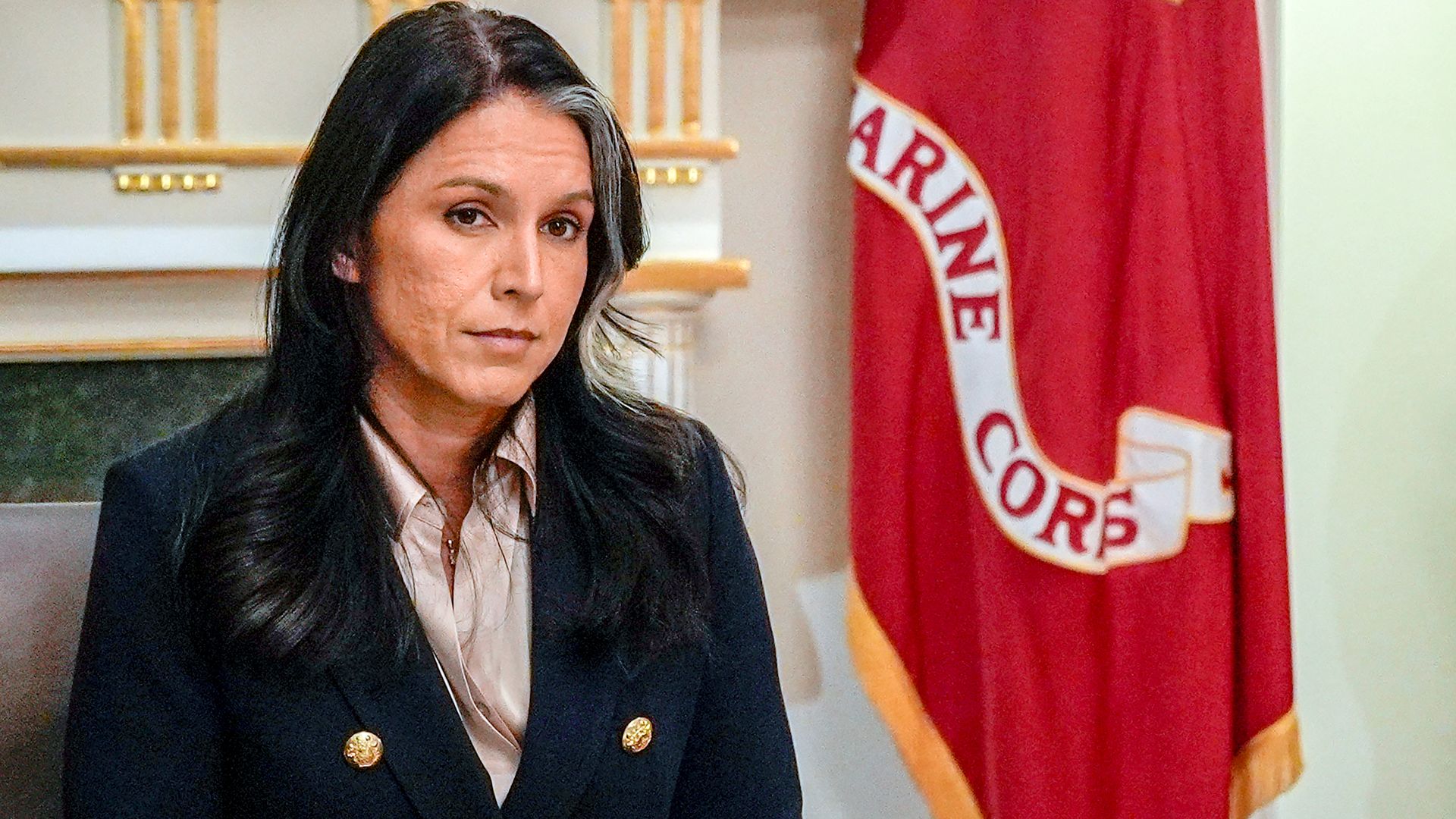 Tulsi Gabbard, who has dark hair with a light face-framing streak, sits in a meeting while wearing a dark jacket with gold buttons and a light collared shirt.