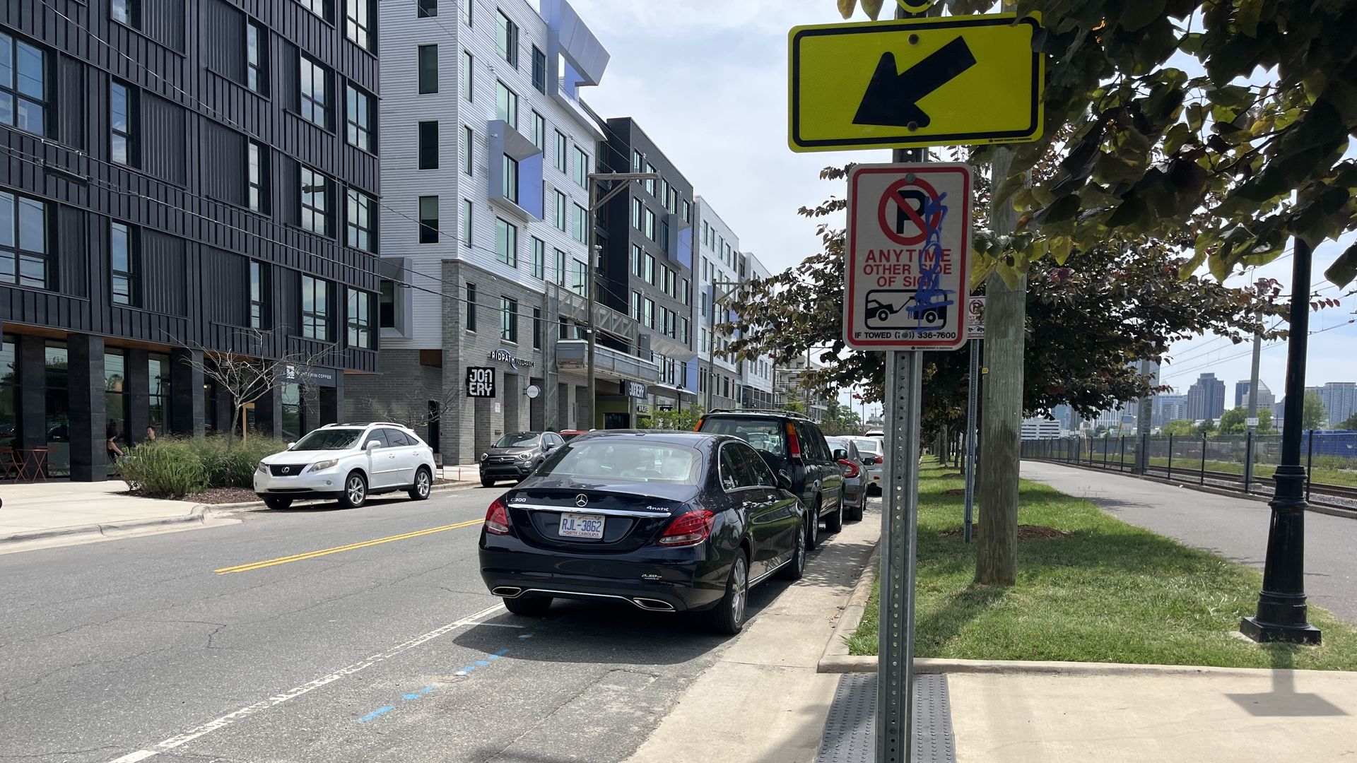 Street scene of a modern urban block with glass-front buildings on the left, several parked cars along the curb, a bright yellow arrow sign and a tow-no parking sign on a post, and trees with a distant skyline.
