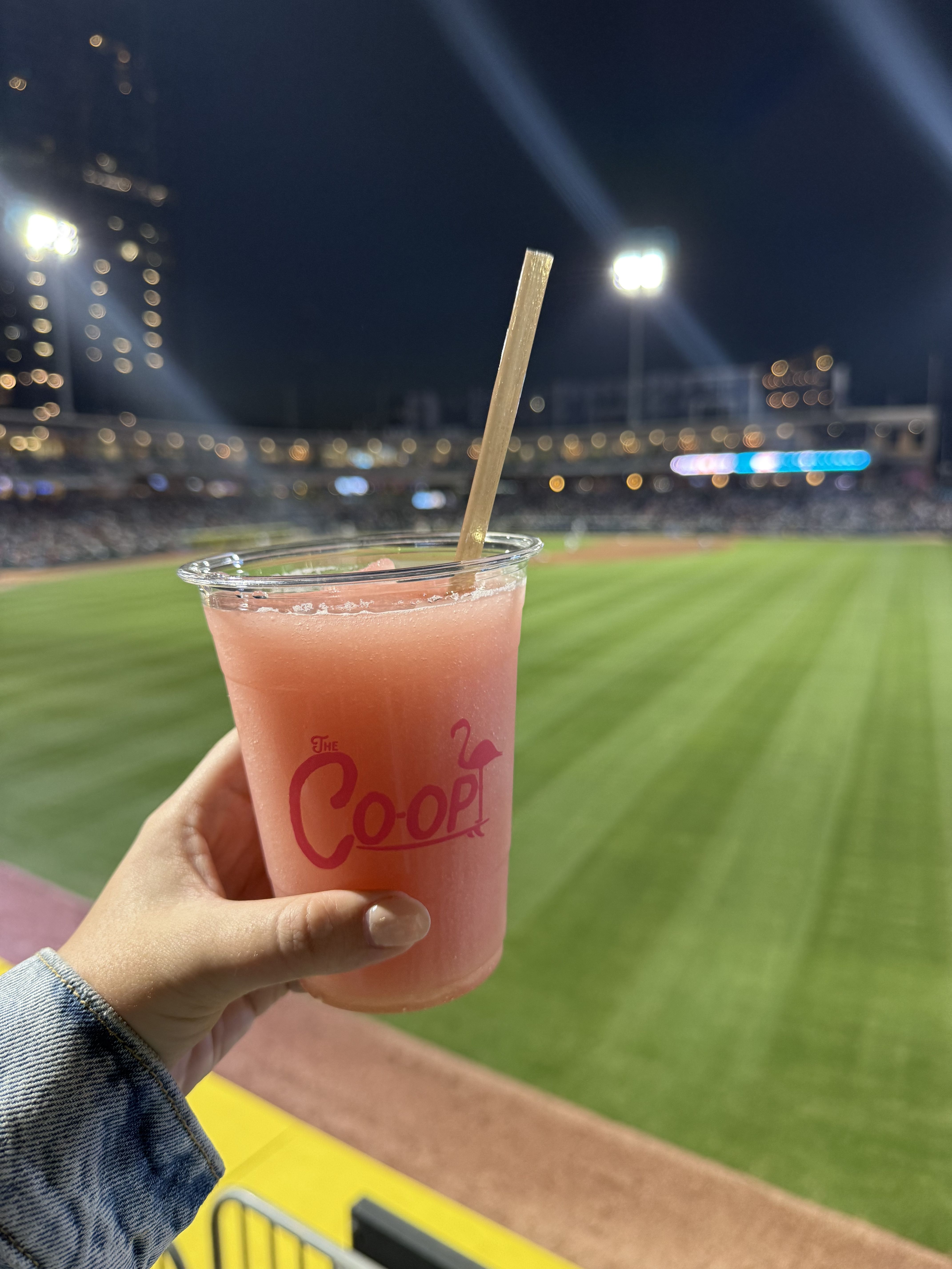 A hand holds a pink frozen drink labeled "The Co-op" with the outfield and night stadium lights in the background.