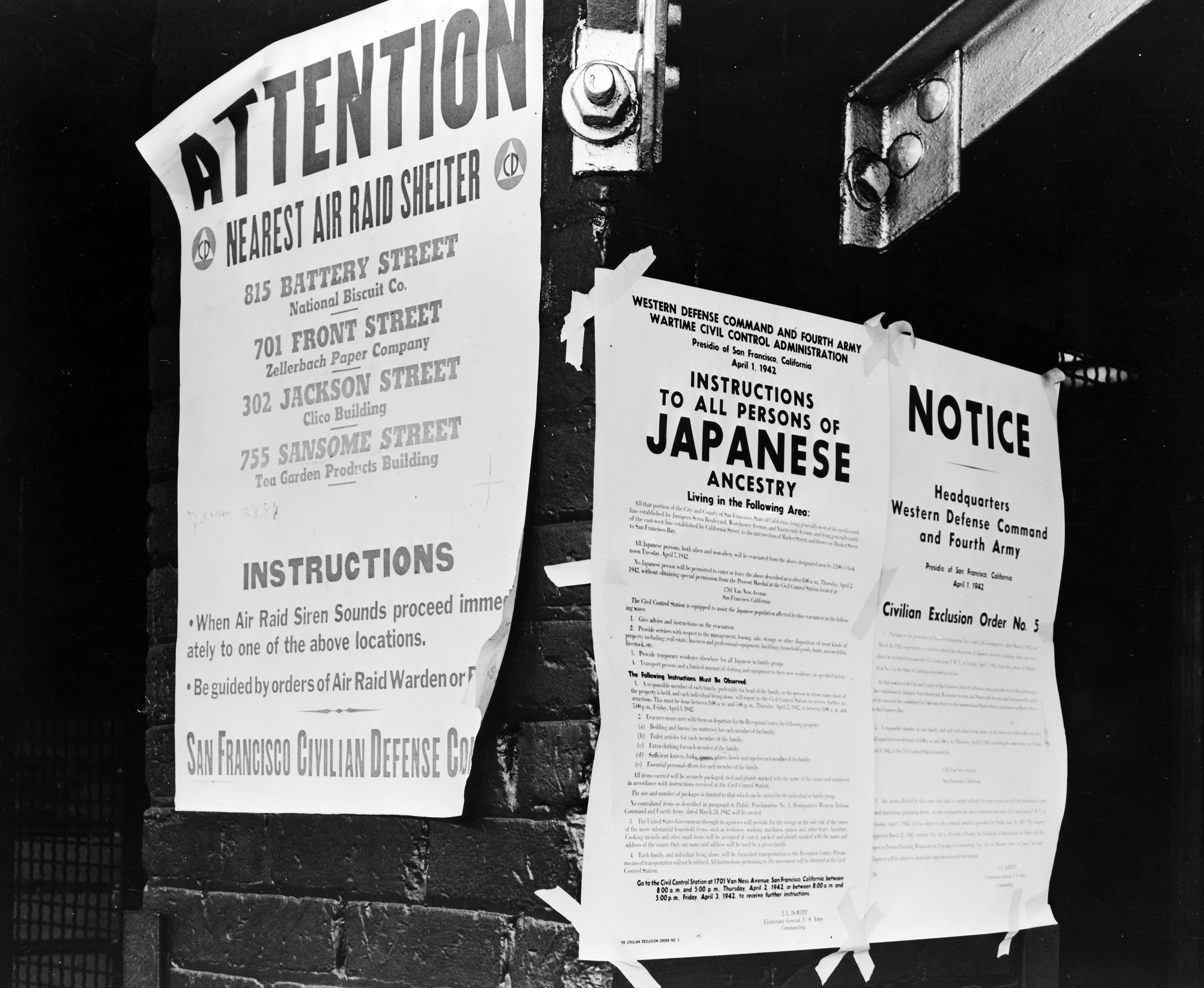 Black-and-white photo of posters pasted on a brick wall, with bold headers like "ATTENTION" and "INSTRUCTIONS" about civil defense; visible bolts and a dark background.