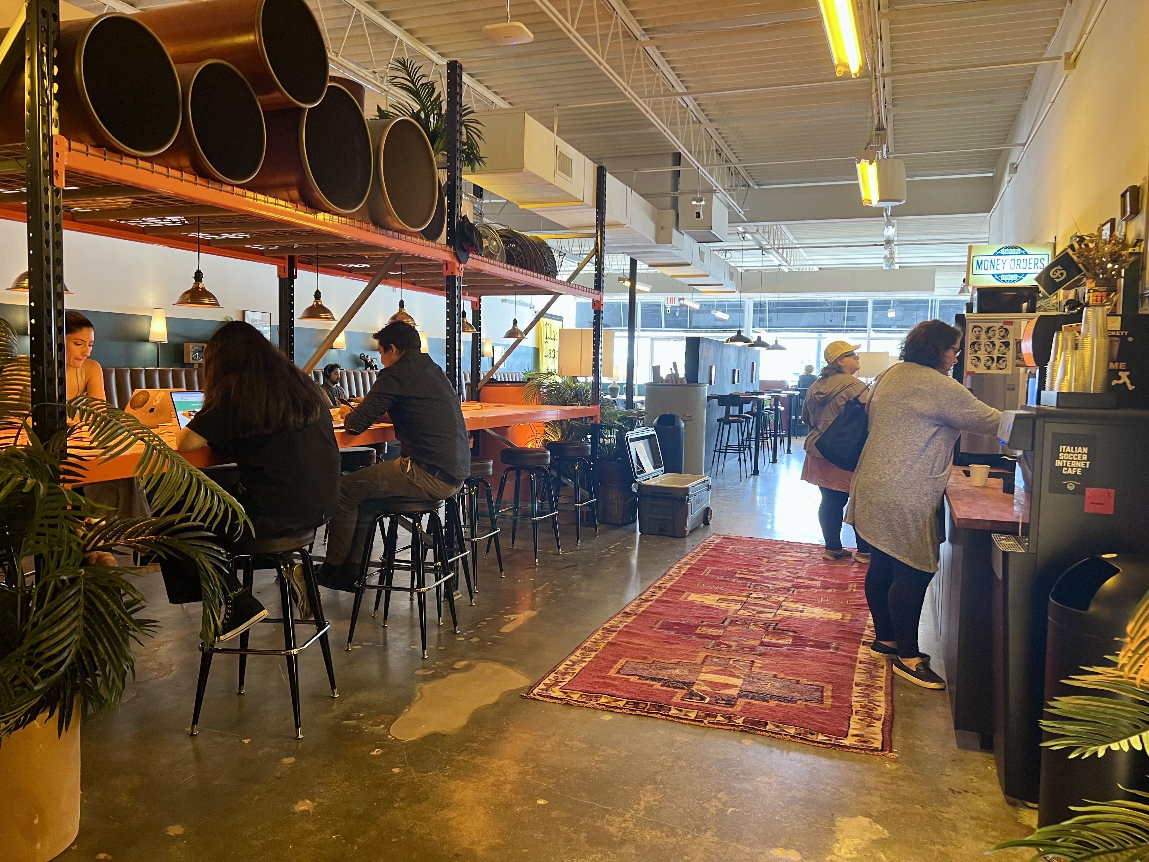 Indoor cafe with people seated on stools at long tables working on laptops, a red patterned rug on the floor, and customers near a counter labeled "Italian Soccer Internet Cafe" and "Money Orders".