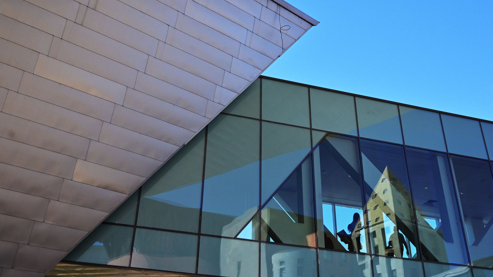 Modern building with an angled metal roof and a glass facade. A blue sky, reflections of a tall nearby building, and silhouettes of people inside visible through the glass.