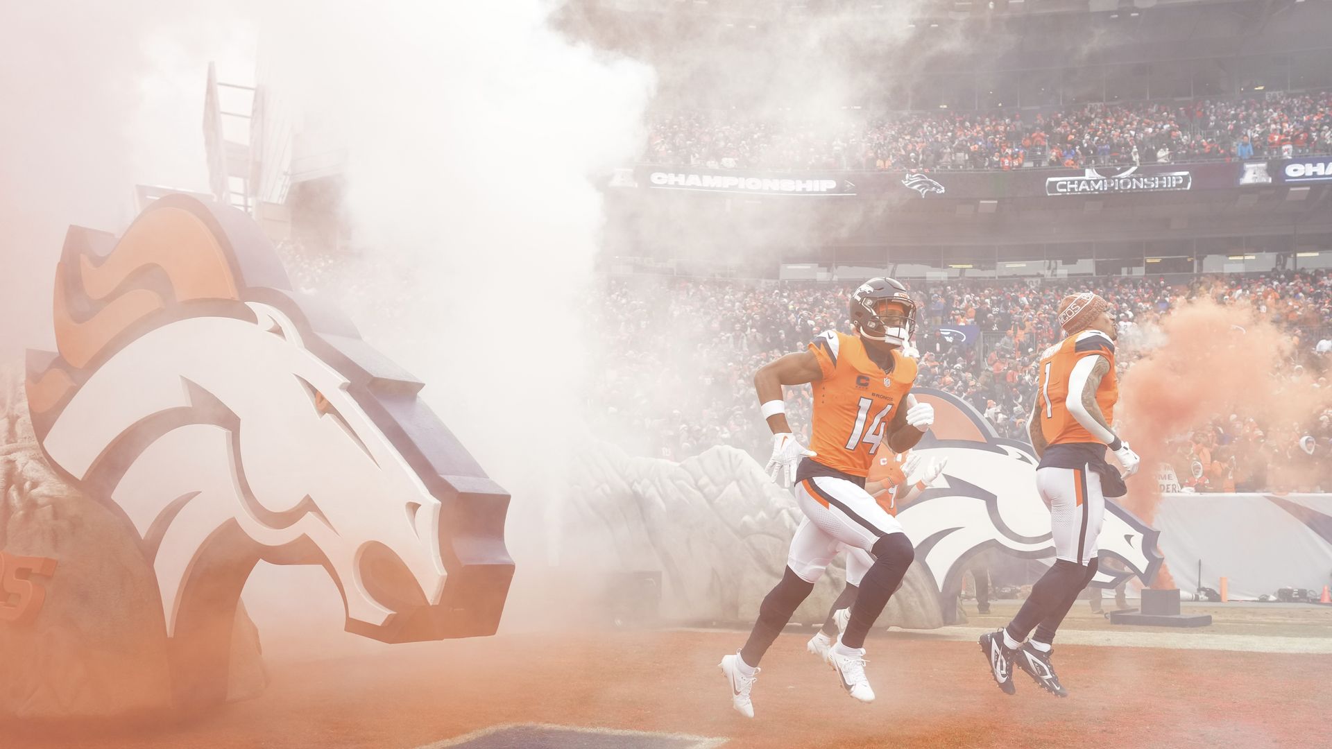 Broncos football stadium scene with a large orange smoke display, a stylized Broncos logo on the left, and players in orange jerseys running onto the field as a crowded stands cheer.