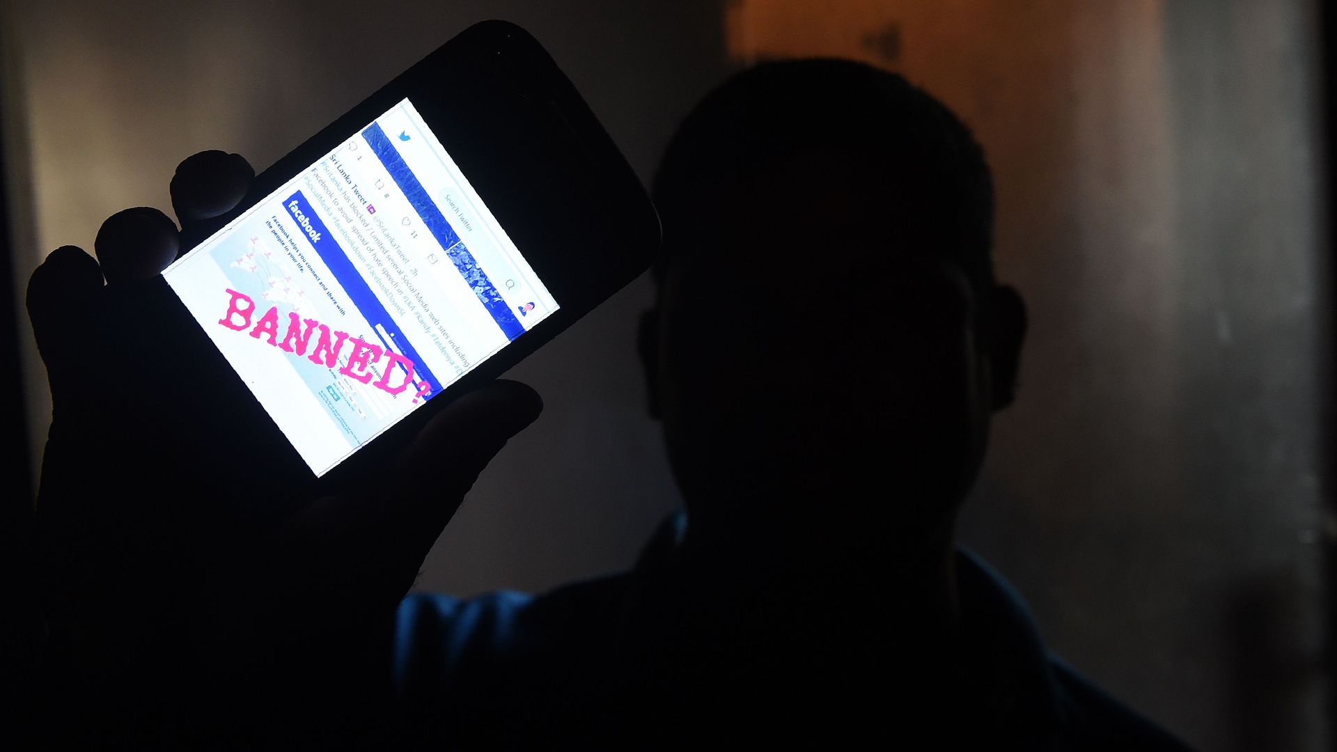 A man holds up a phone showing the Facebook app and "BANNED" written across the screen