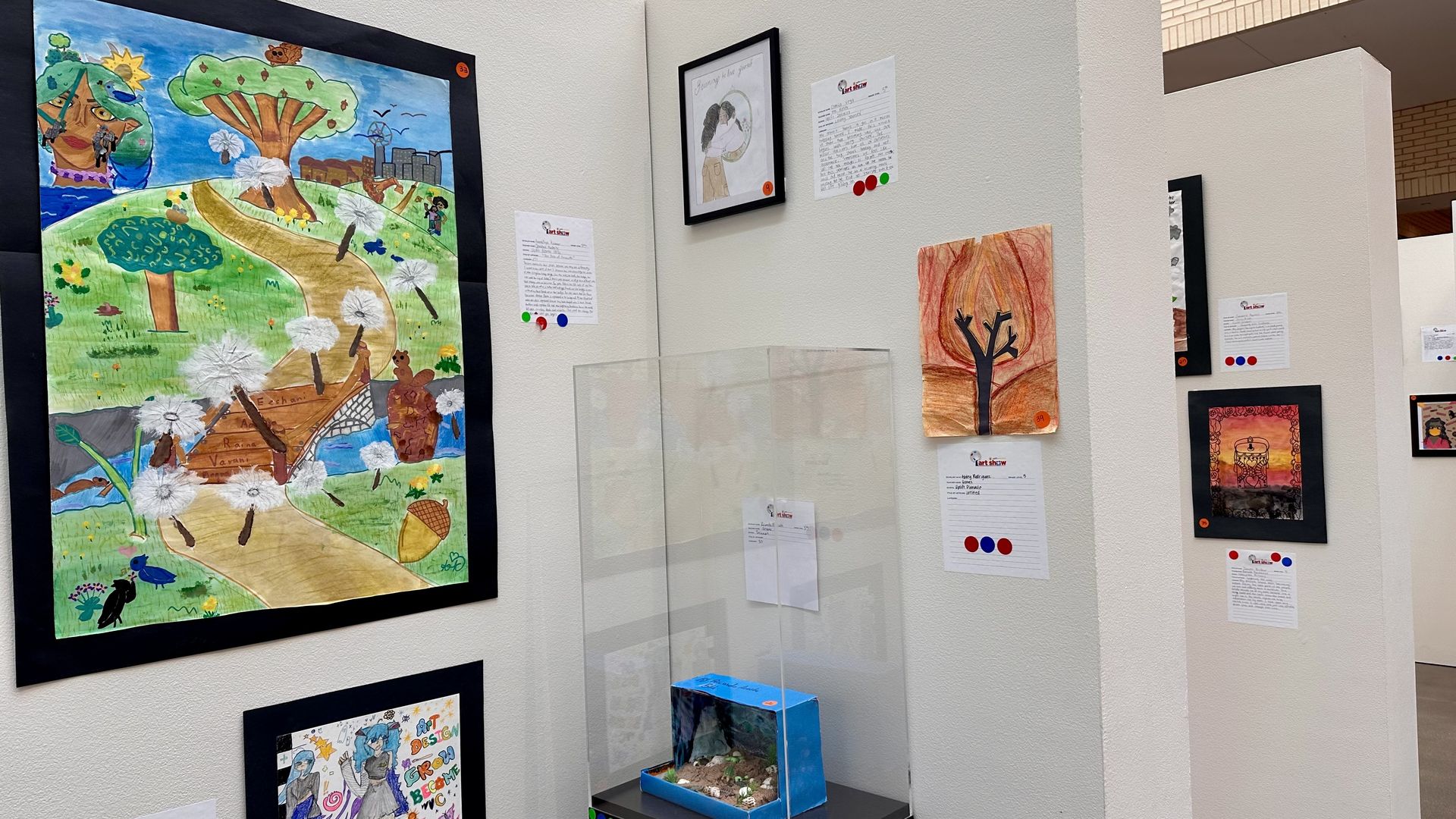 Art exhibit featuring colorful children's drawings of nature and abstract scenes, displayed on white walls with descriptive cards and red, blue, green voting stickers. One 3D diorama in a clear case.