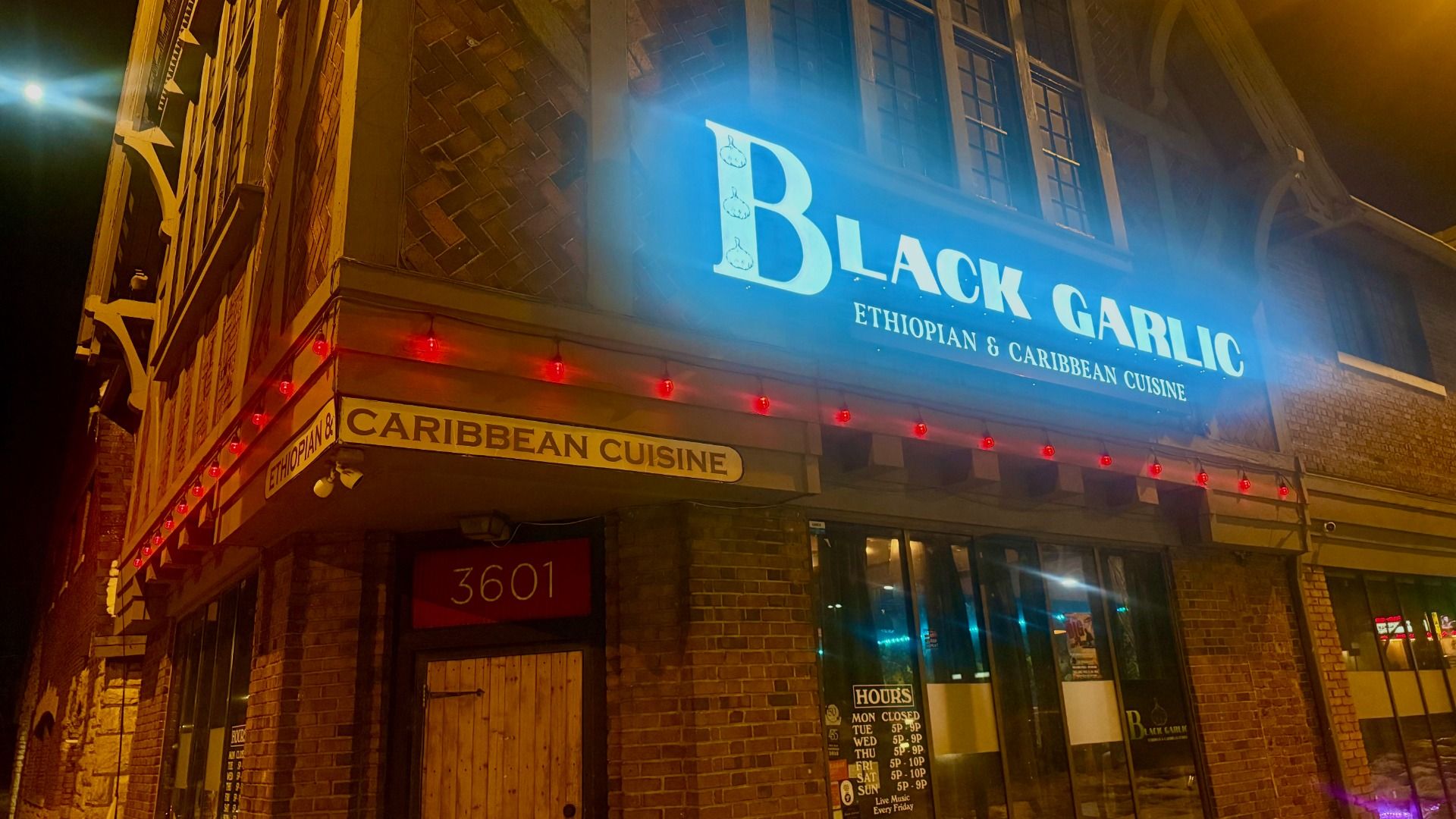 Night view of Black Garlic restaurant with illuminated blue sign offering Ethiopian and Caribbean cuisine, red string lights, brick exterior, and posted hours at entrance.