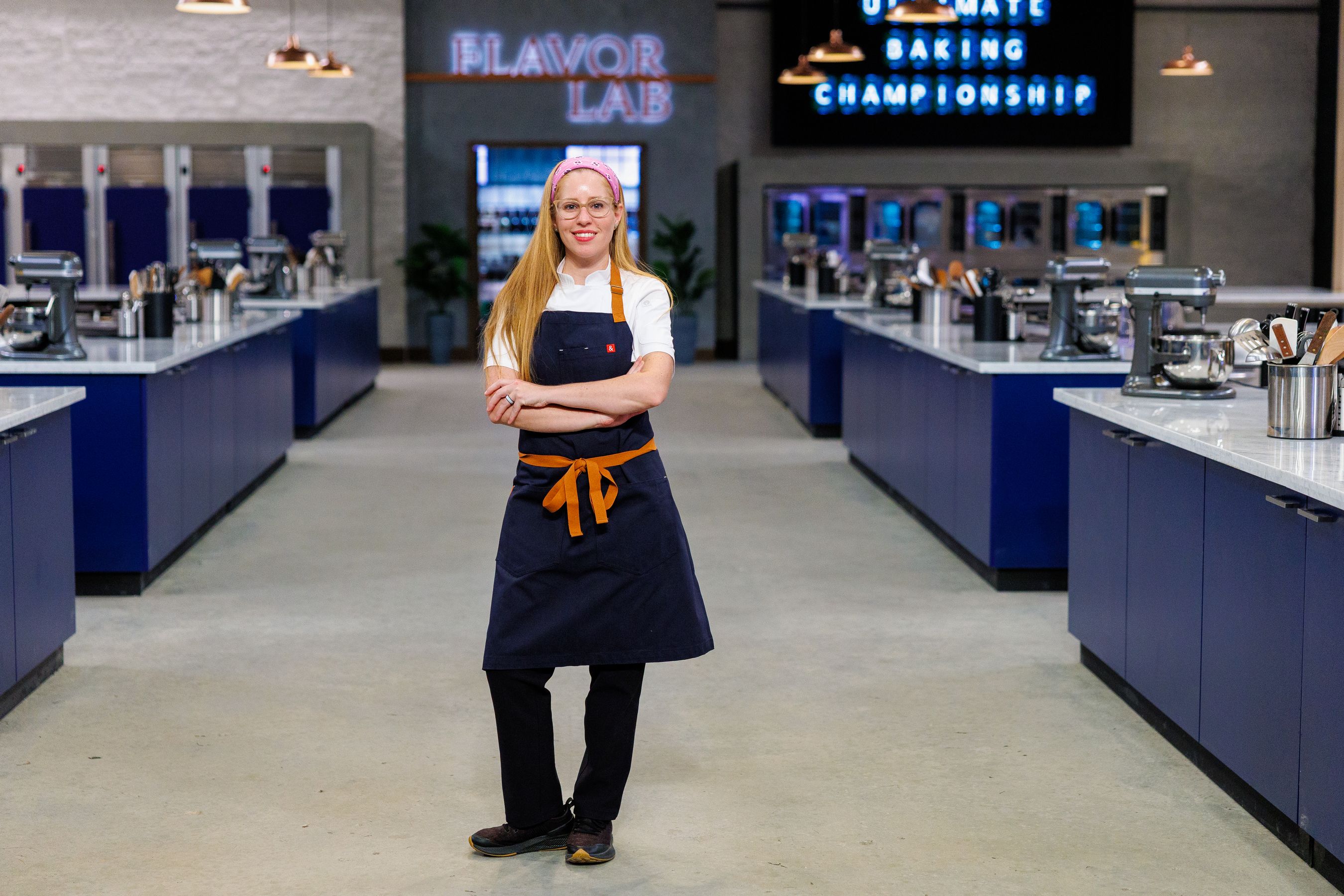 Smiling chef Rochelle Cooper in navy apron with orange tie and pink headband stands with arms crossed in a modern kitchen labeled Flavor Lab for the Ultimate Baking Championship.