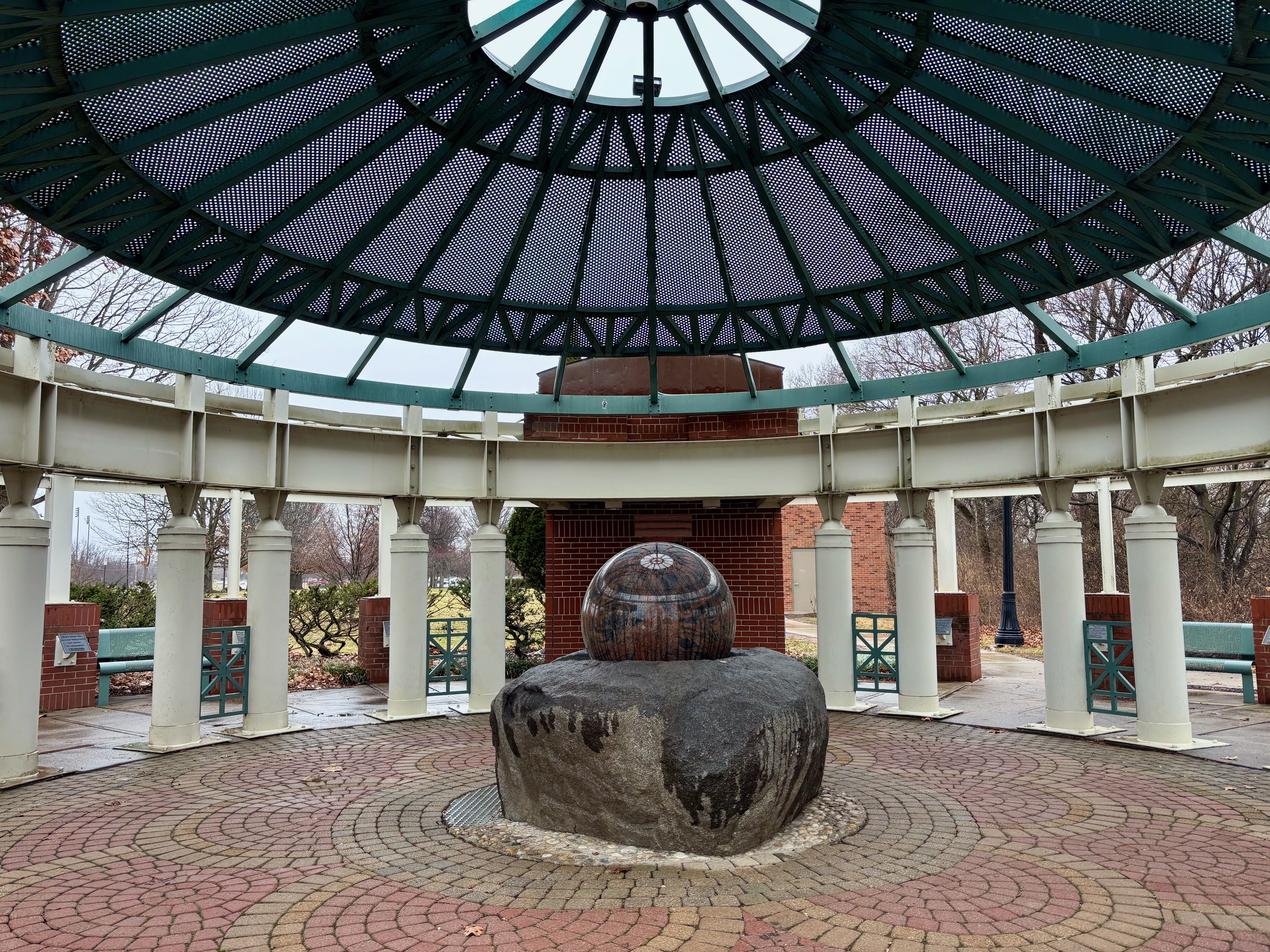 Circular outdoor pavilion with white columns and teal metal roof, featuring a large polished stone sphere on a rough stone base in the center on patterned brick pavement.