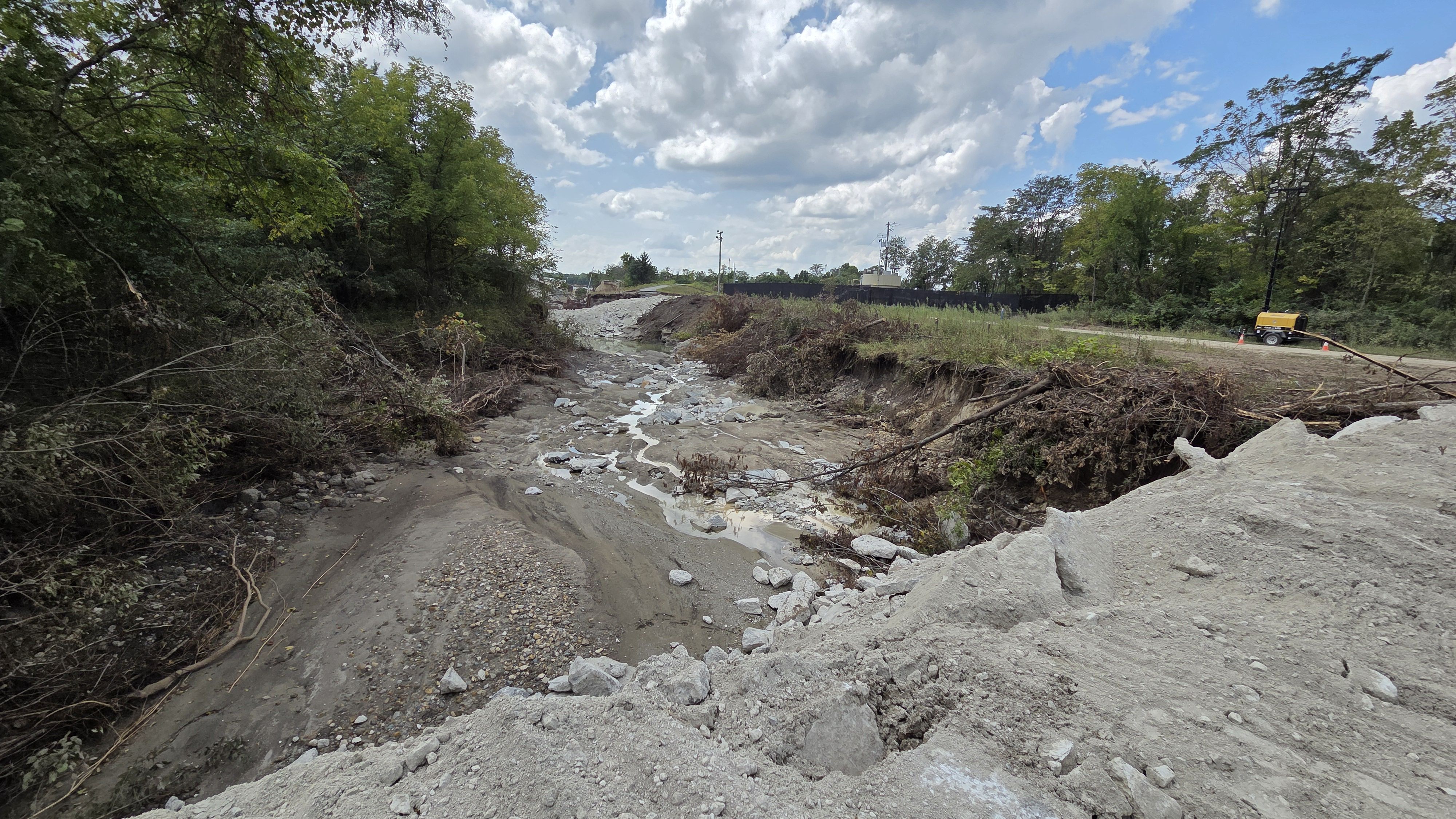Wide construction scene with a rocky, eroded riverbed flanked by trees. A yellow portable generator sits on a dirt road with orange cones; blue sky with fluffy clouds overhead.