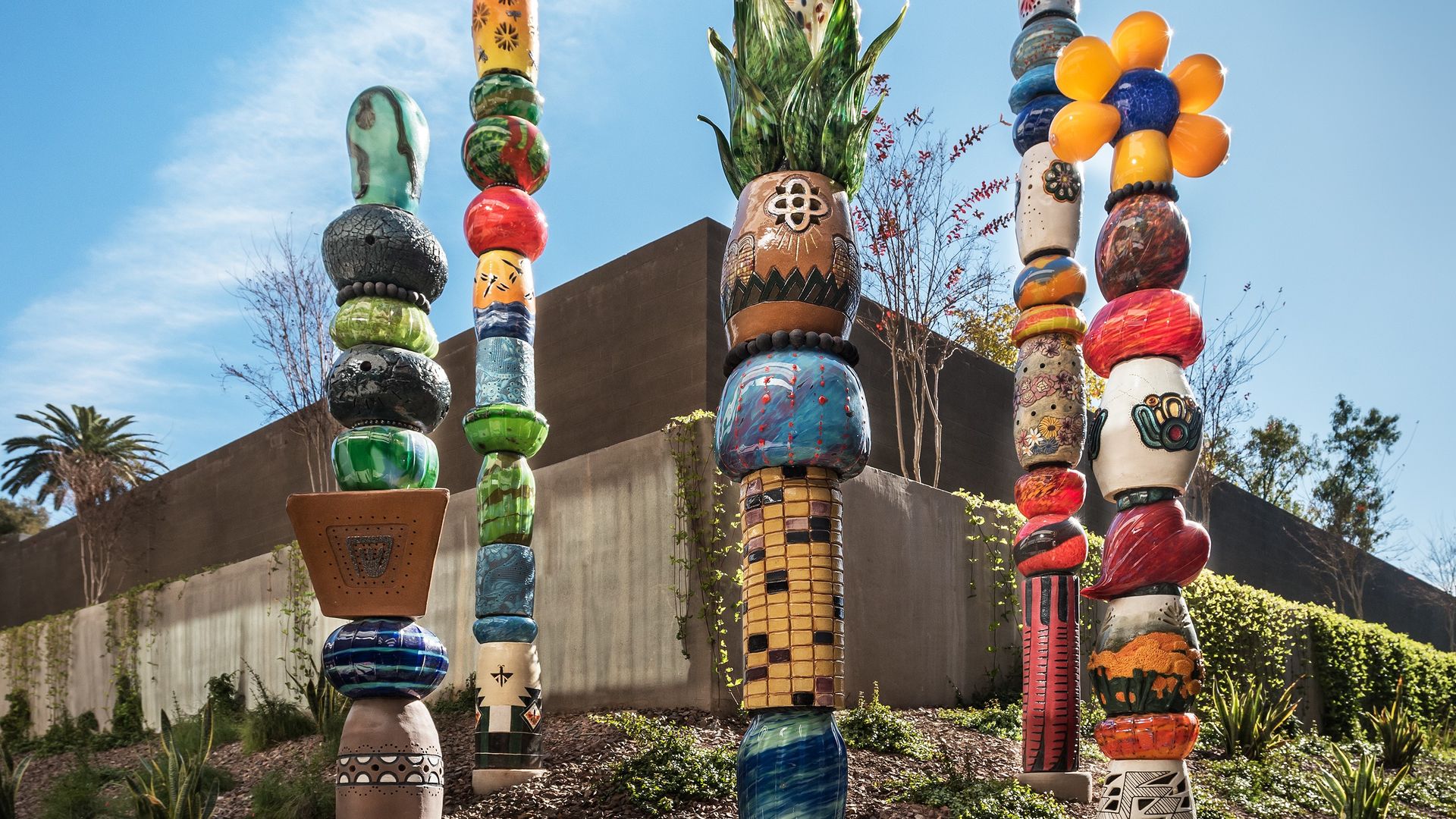 Colorful ceramic totem poles with various patterns and shapes stand outdoors against a blue sky, surrounded by greenery and a concrete wall in the background.