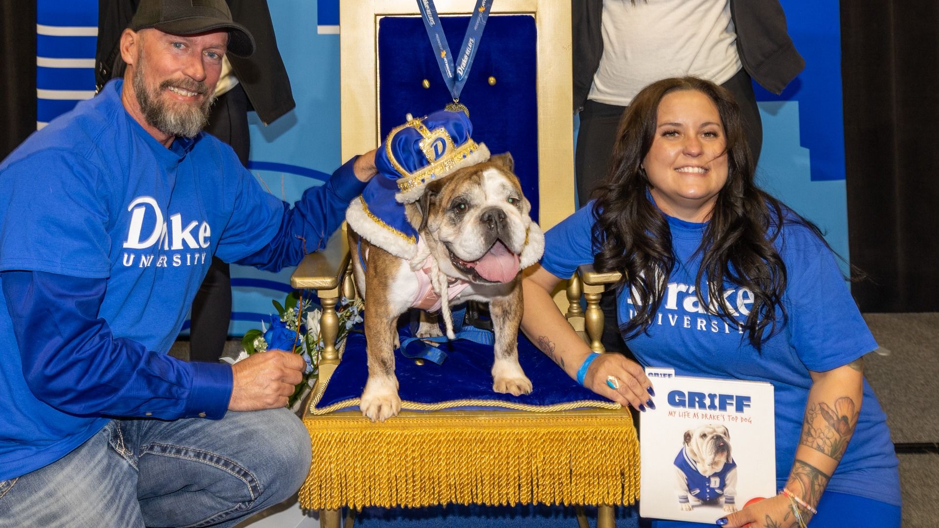 Three people in Duke University blue shirts pose with a bulldog wearing a crown and cape, seated on a blue velvet throne. A "GRIFF" display sits beside them.