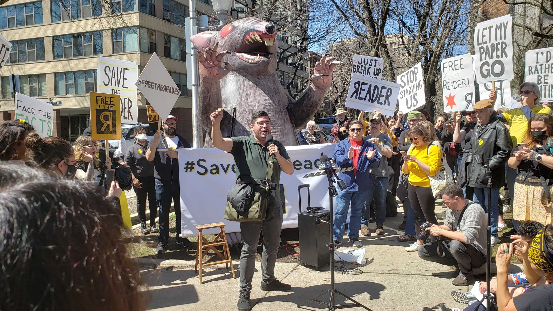 Photo of protesters rallying around a speaker, who stands in front of an inflated rat.