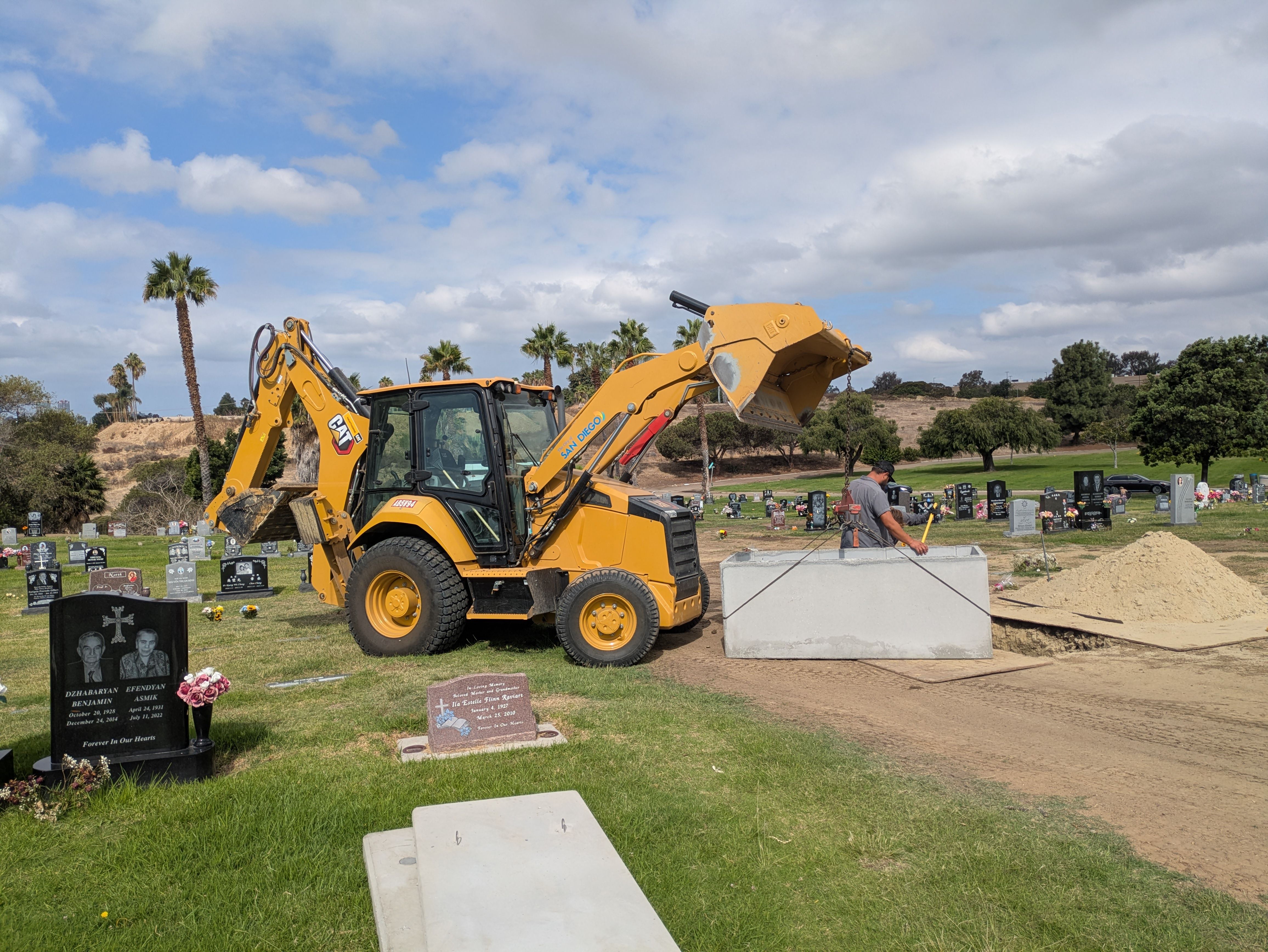 Bright yellow backhoe excavator sits at a cemetery, surrounded by headstones and green grass. A worker inspects a dirt mound; palm trees dot the background beneath a partly cloudy blue sky.