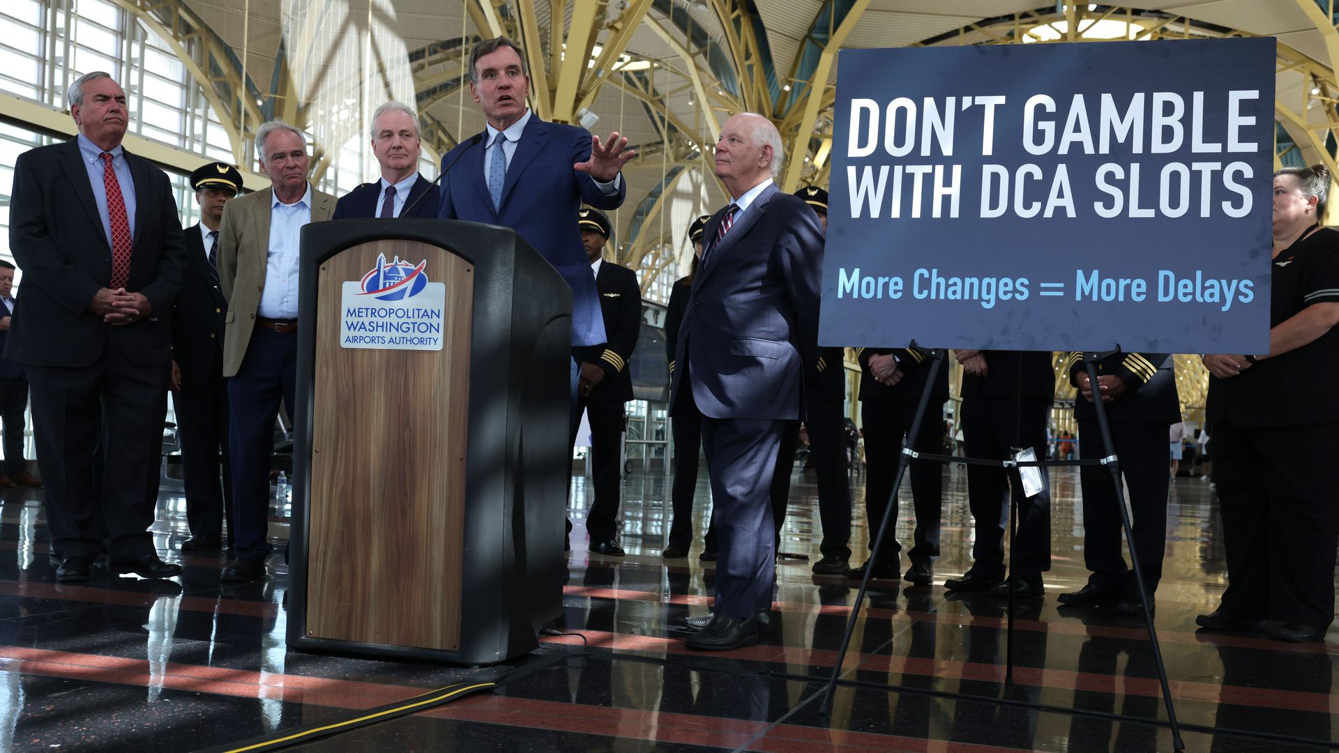 Sen Mark Warner speaks at a podium with a sign that says "Don't Gamble with DCA Slots" and "More Changes = More Delays"