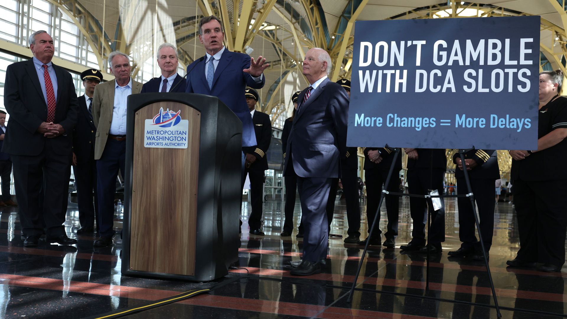 Sen Mark Warner speaks at a podium with a sign that says "Don't Gamble with DCA Slots" and "More Changes = More Delays"