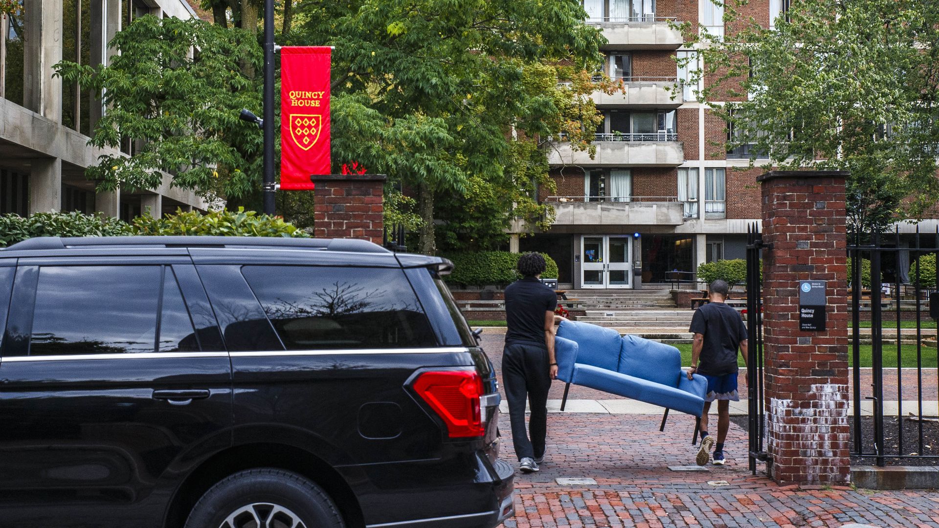 Two people carry a couch and walk it past a gate. A black car is behind them, but in the foreground of the photo. Trees are seen outside the brick building 