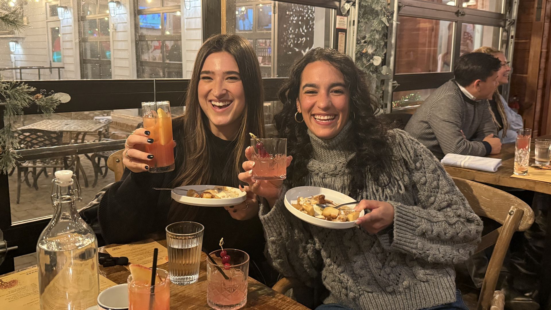 Two women smiling at a wooden table in a warmly lit restaurant, holding drinks and plates of dessert, with festive decorations and other diners in the background.