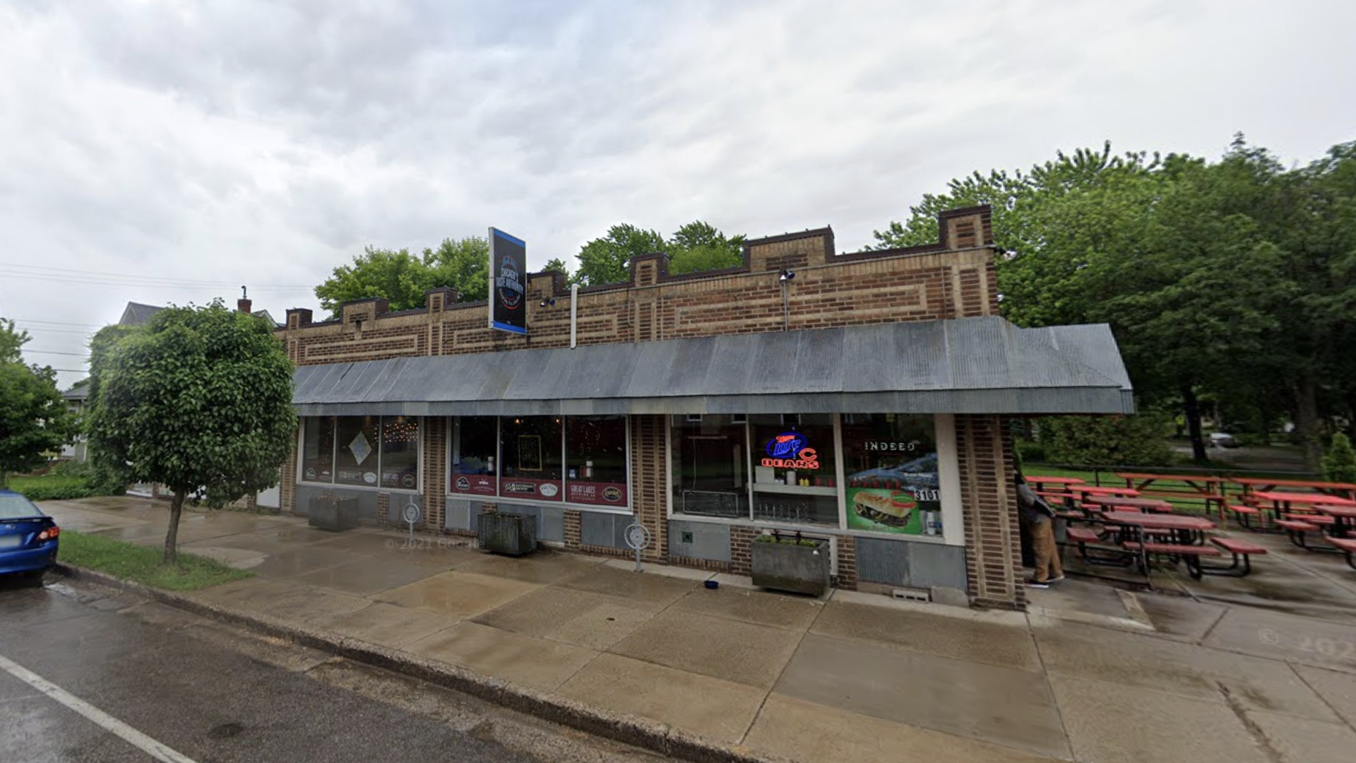 Brick building with large windows and a metal awning houses a business with a blue sign. Red picnic tables and green trees are visible outside on a wet sidewalk under a cloudy sky.