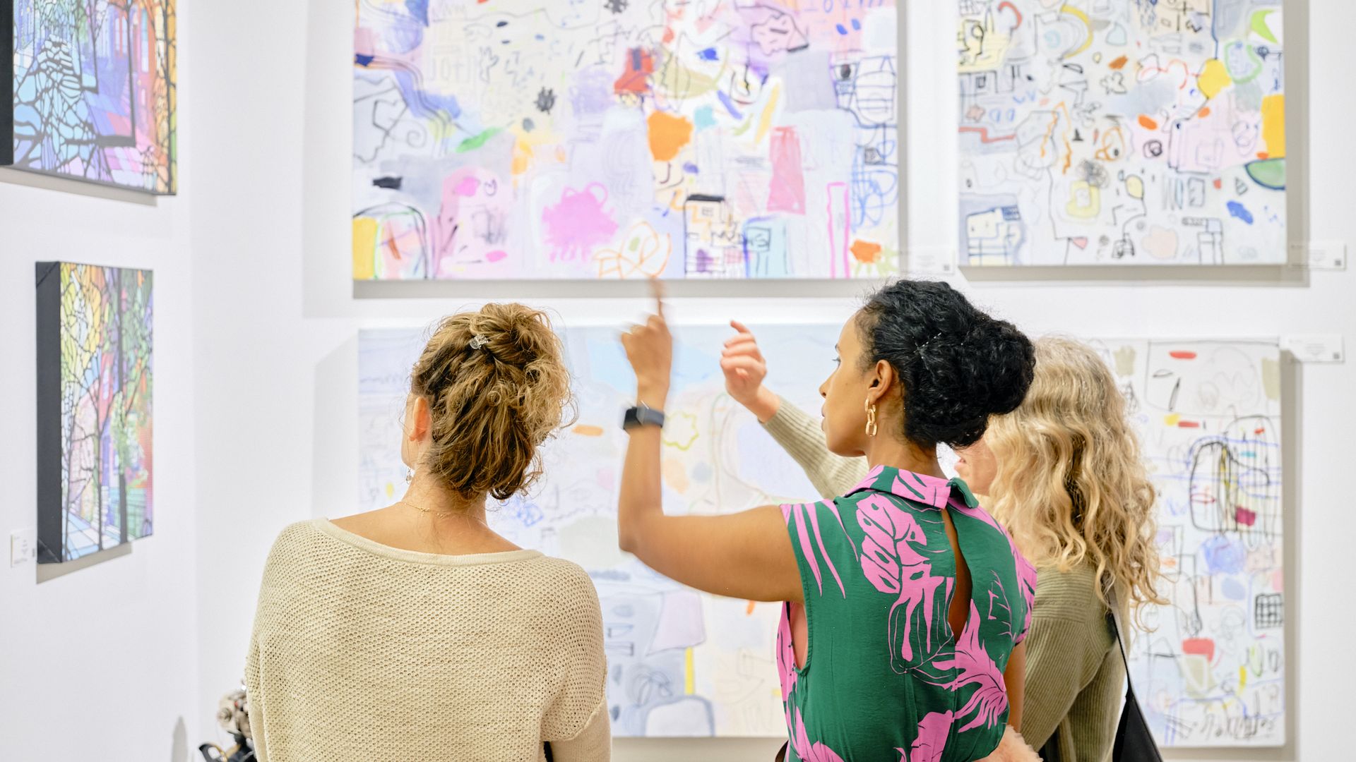 Three women look at artwork on the corner of a room as the woman in the middle points at the artwork.