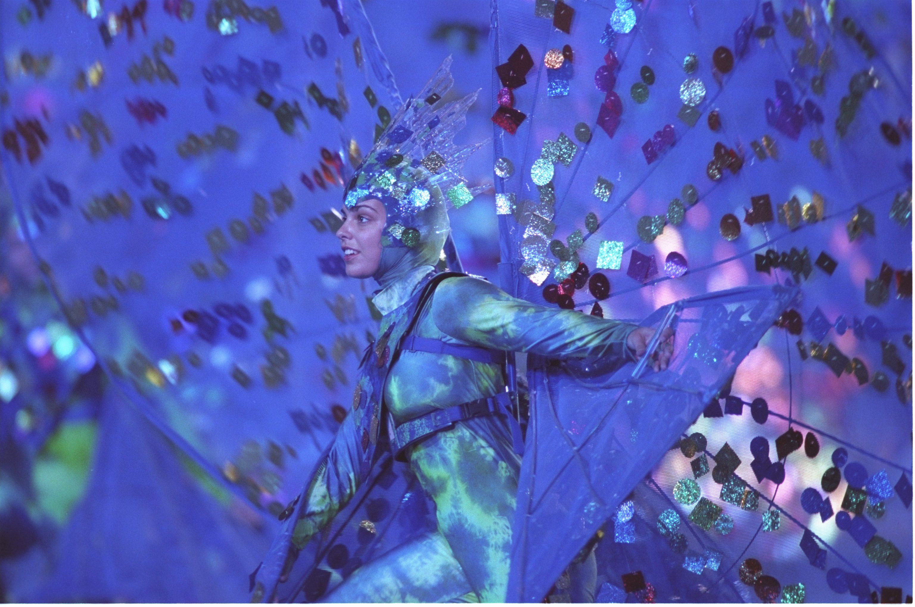 A dancer in a full body blue costume with giant peacock-like wings performs