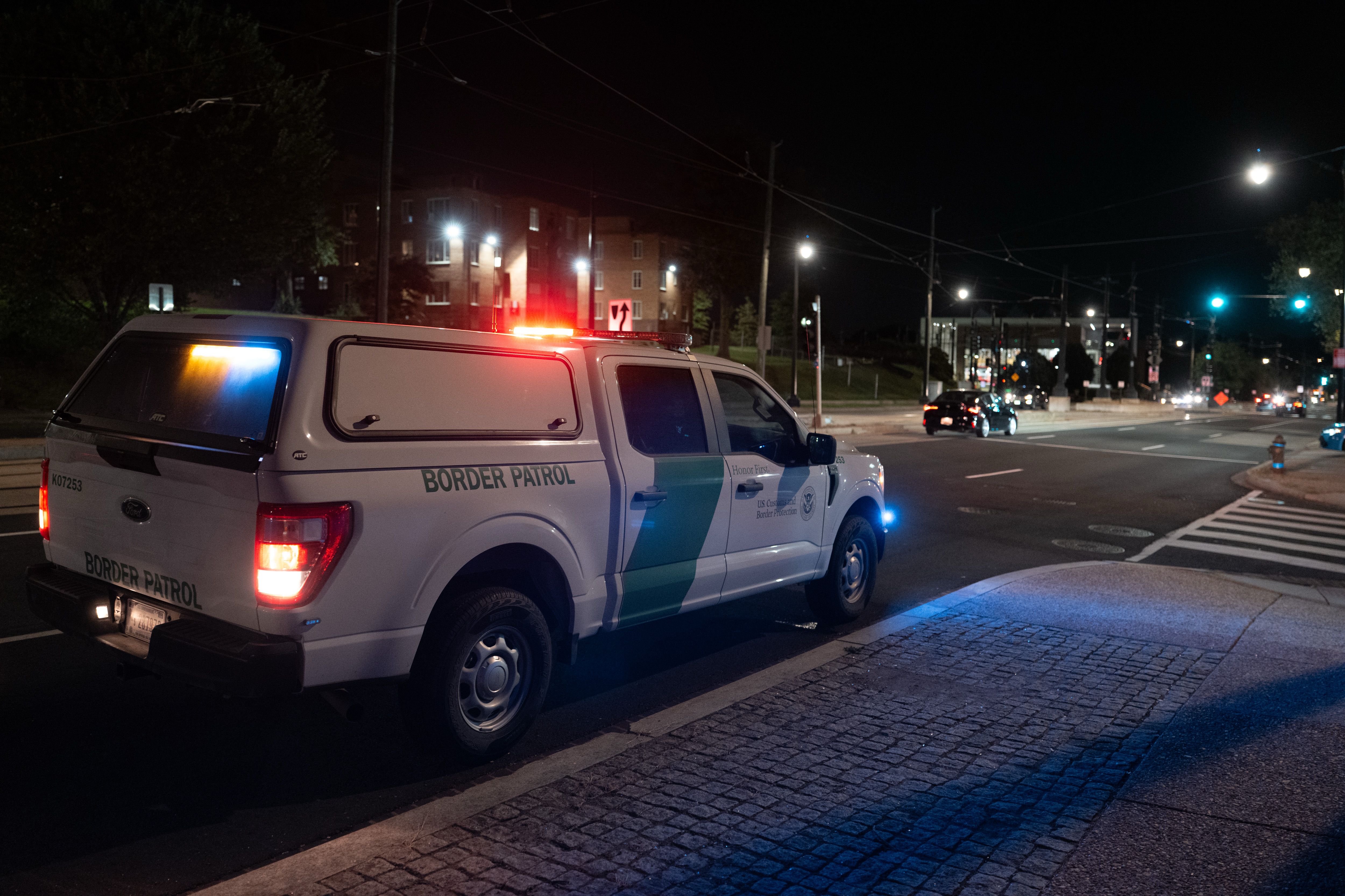 White U.S. Border Patrol truck with red and blue lights on, parked on a street at night near a crosswalk and traffic lights in an urban area.