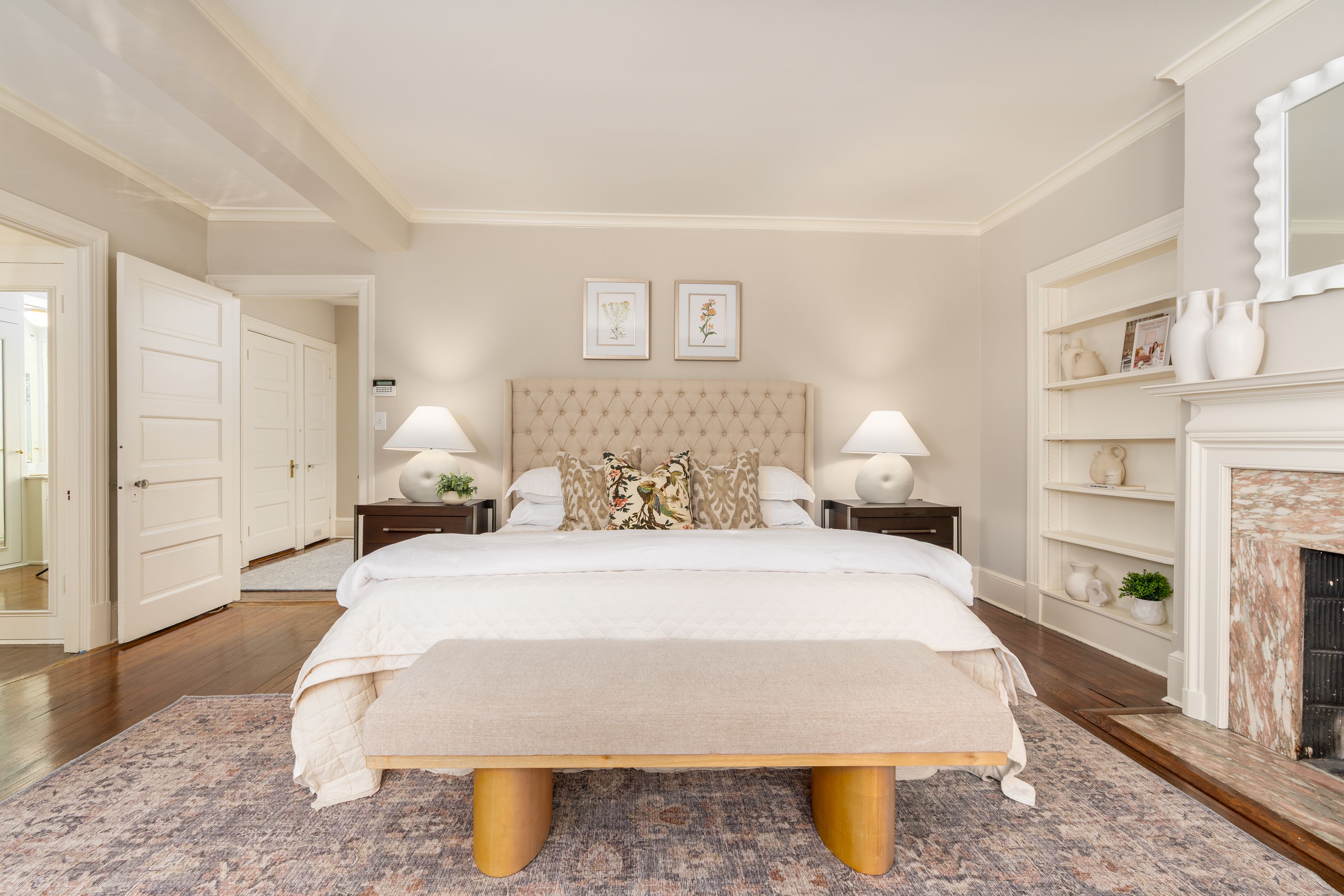 Bright beige bedroom with a tufted beige headboard, two nightstands and lamps, botanical prints above bed, white doors left, built-in shelves and white vases, marble fireplace on right.