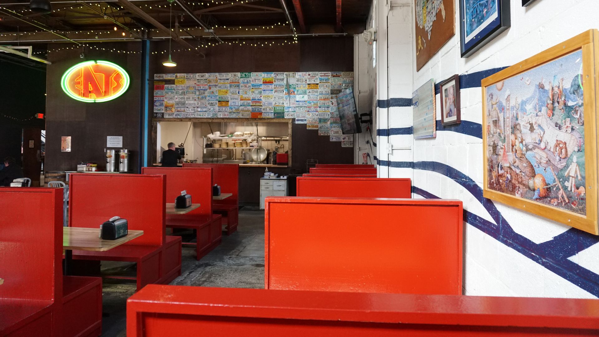 Interior of a casual diner with bright red booths, white walls with blue stripes, string lights, and an open kitchen in back. A license-plate mosaic wall and framed art line the walls; a neon sign glows on the left.