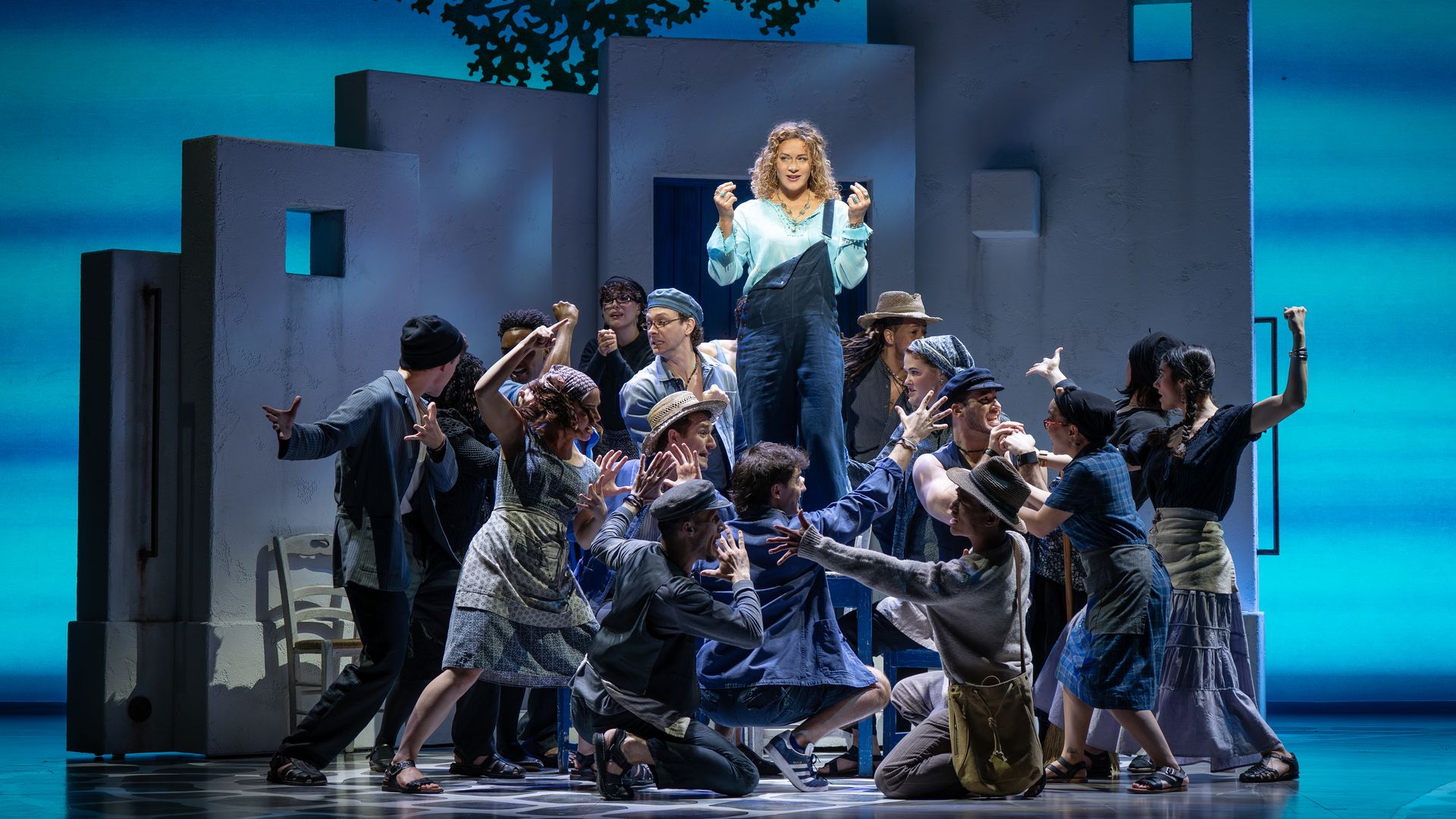 Stage performance: a woman in a light blue top and overalls stands on a chair, surrounded by dancers in casual clothes, against a gray block set and a blue gradient backdrop.